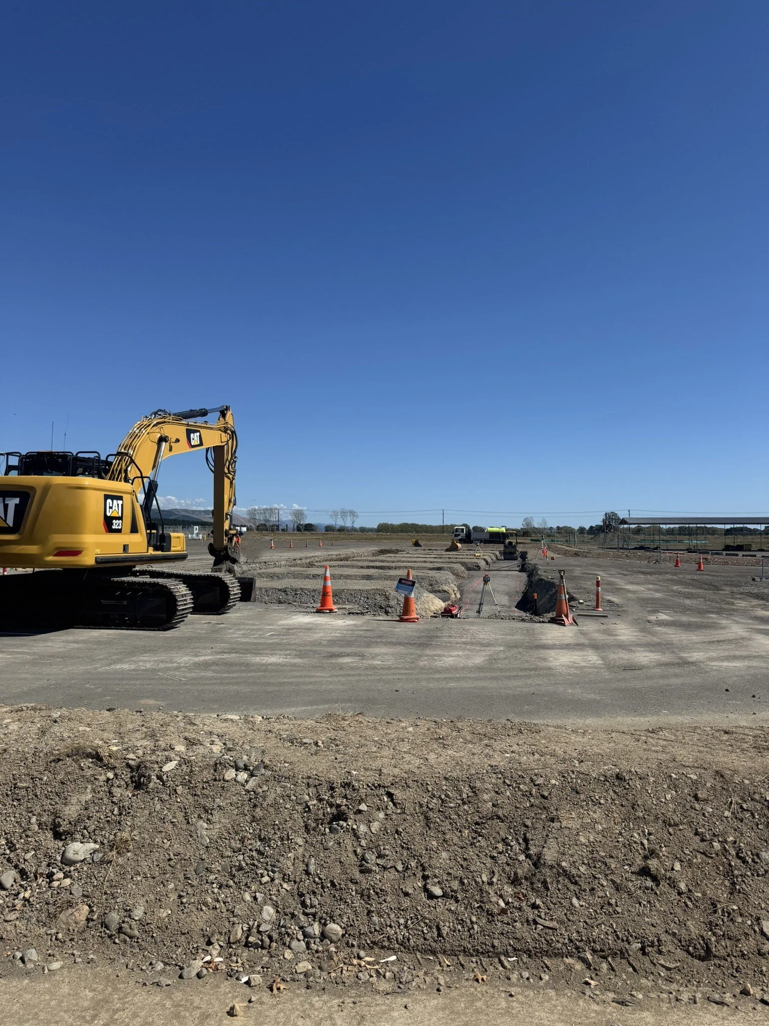 Construction site with a large yellow Caterpillar excavator, orange safety cones, and machinery working on the dirt ground under a clear blue sky.
