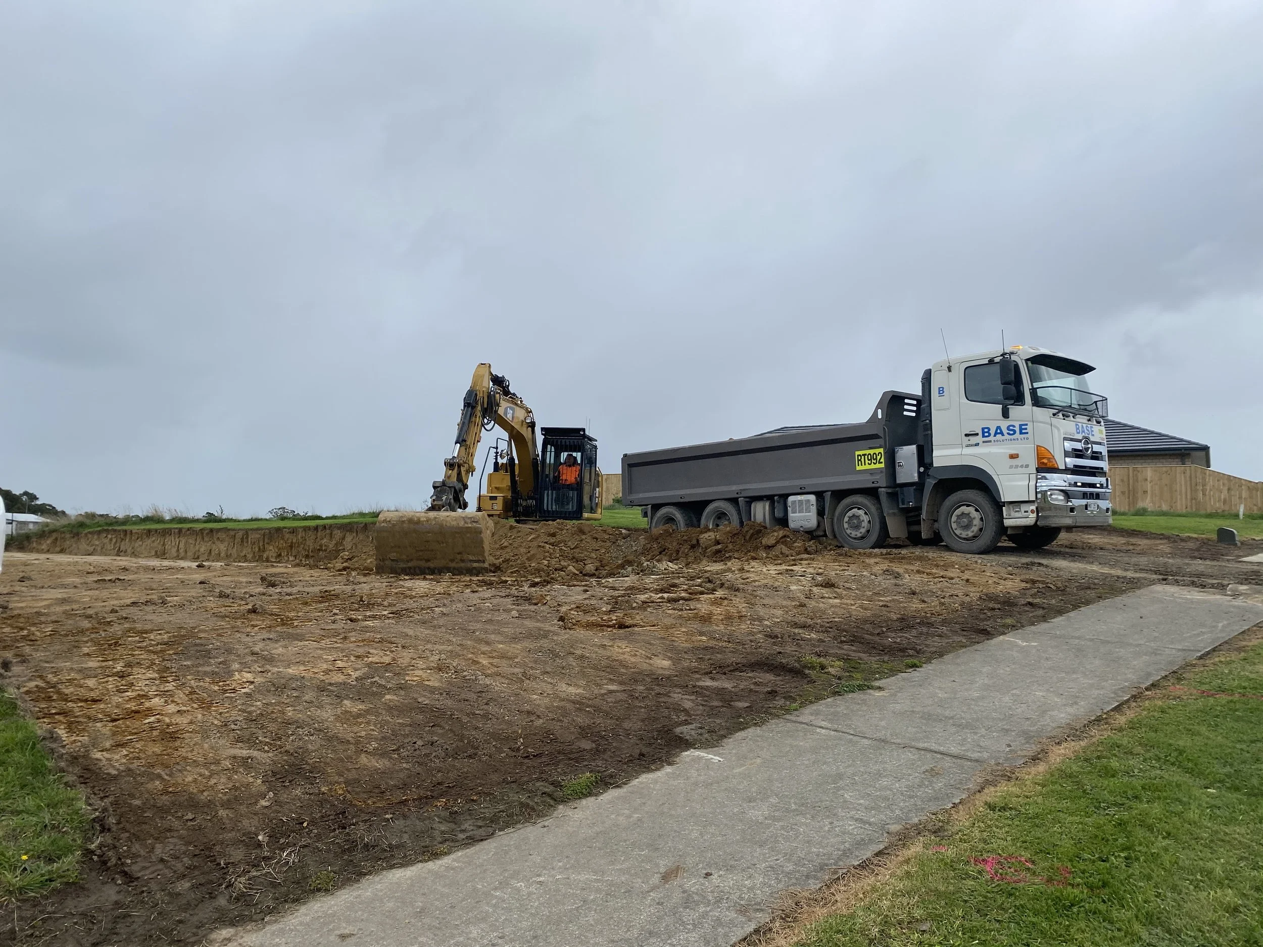 Construction site with a yellow excavator and a white dump truck on dirt ground, overcast sky.