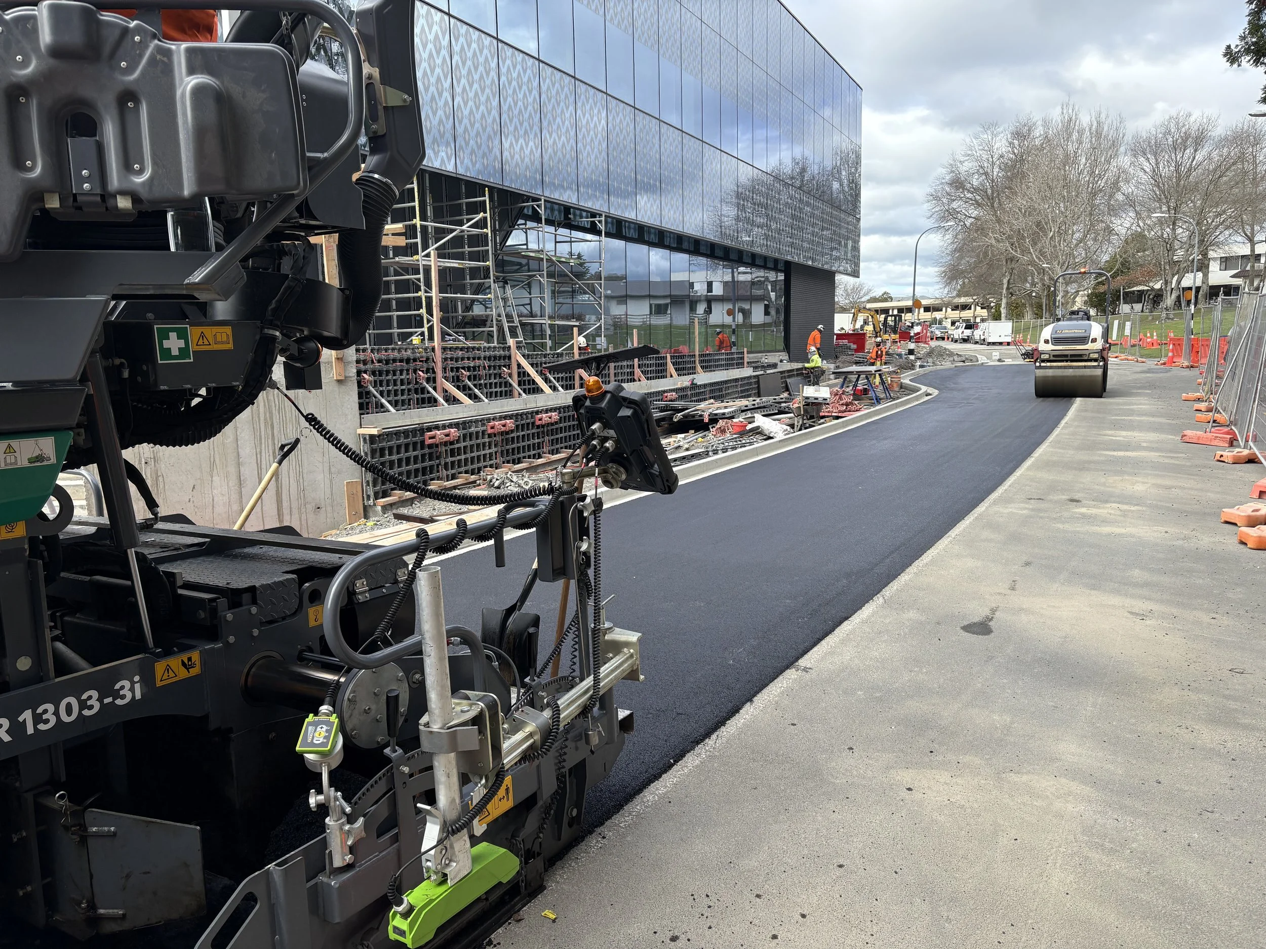 Construction workers paving a curved street with asphalt, with a roller compactor rolling the new pavement. Building scaffolding and fencing are visible along the sidewalk, and a modern glass building is in the background.