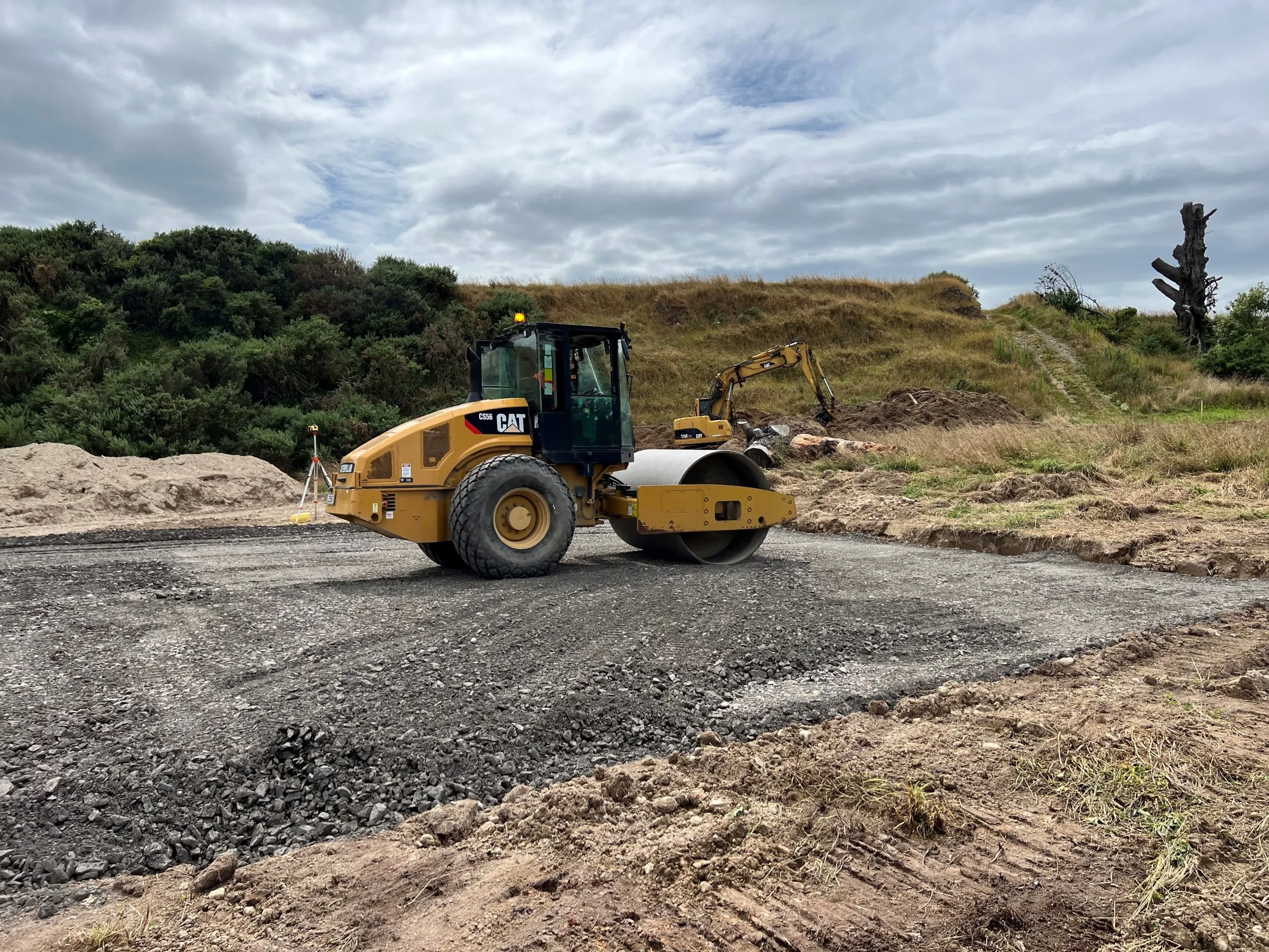 Construction site with a yellow Caterpillar road roller and a mini excavator working on a dirt hillside under a cloudy sky.