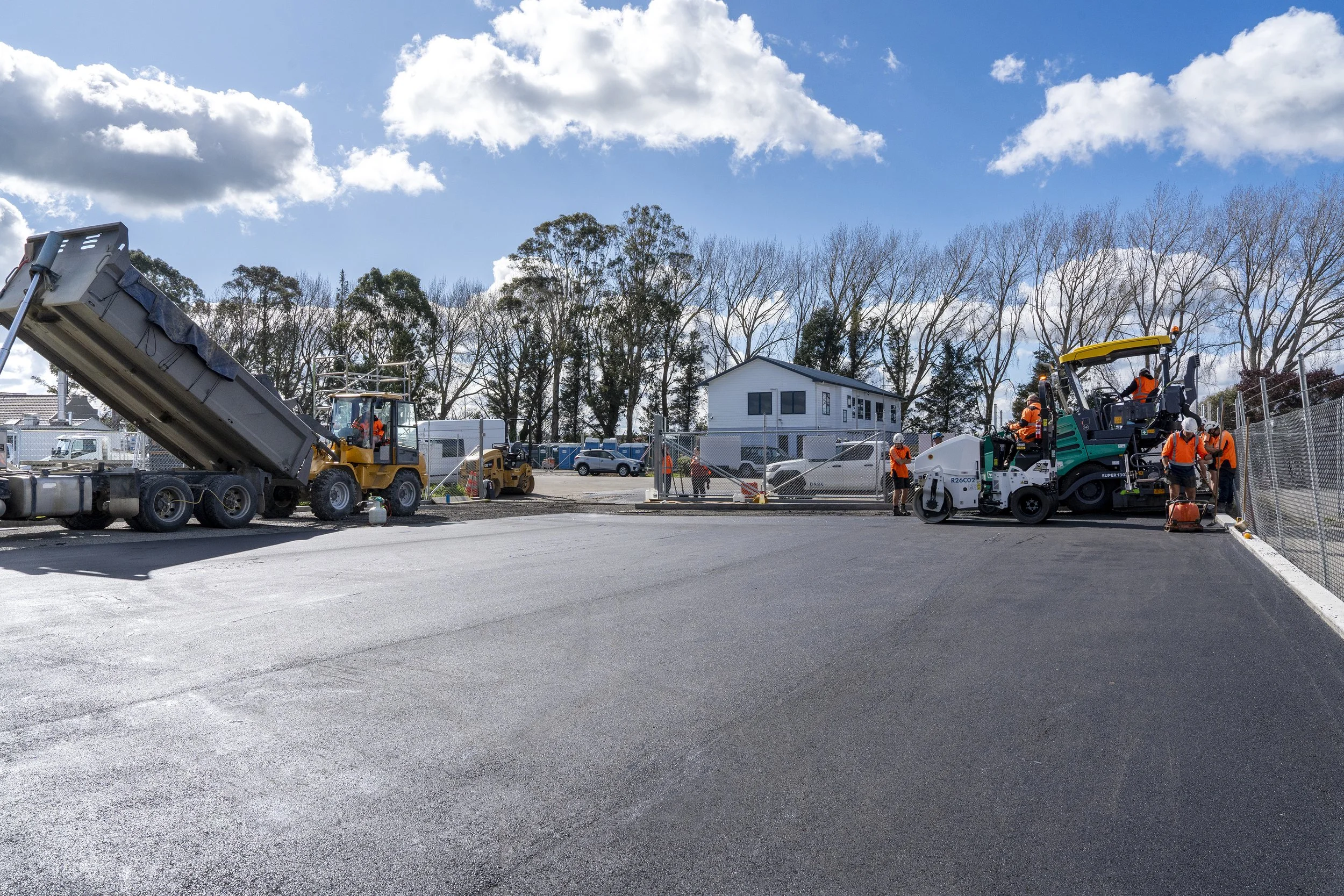 Construction workers paving a road with asphalt, operating heavy machinery under a partly cloudy sky, with a house and trees in the background.