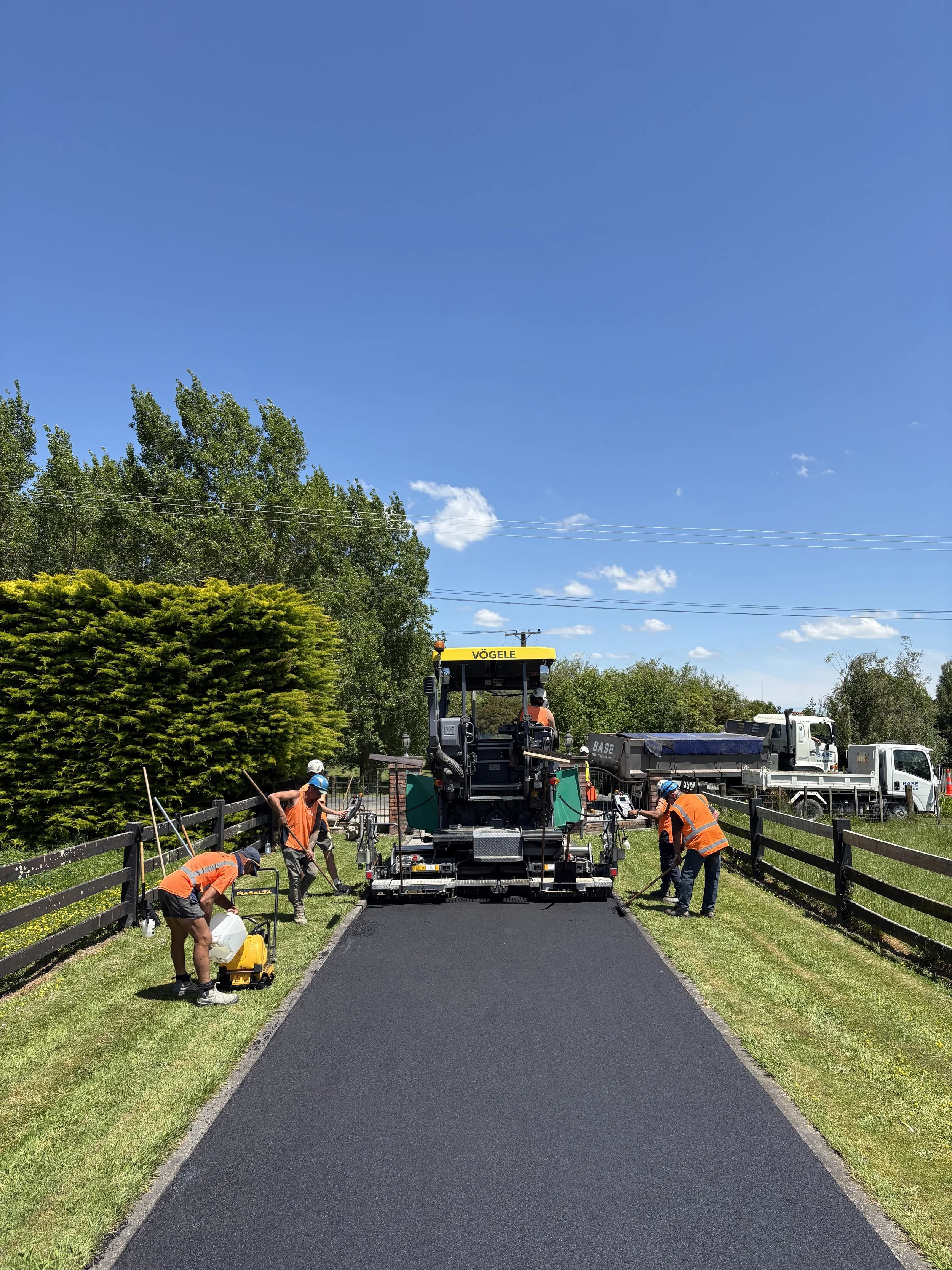 Workers in orange safety vests and helmets laying fresh asphalt on a pathway, flanked by wooden fences, with trees and trucks in the background under a blue sky with a few clouds.