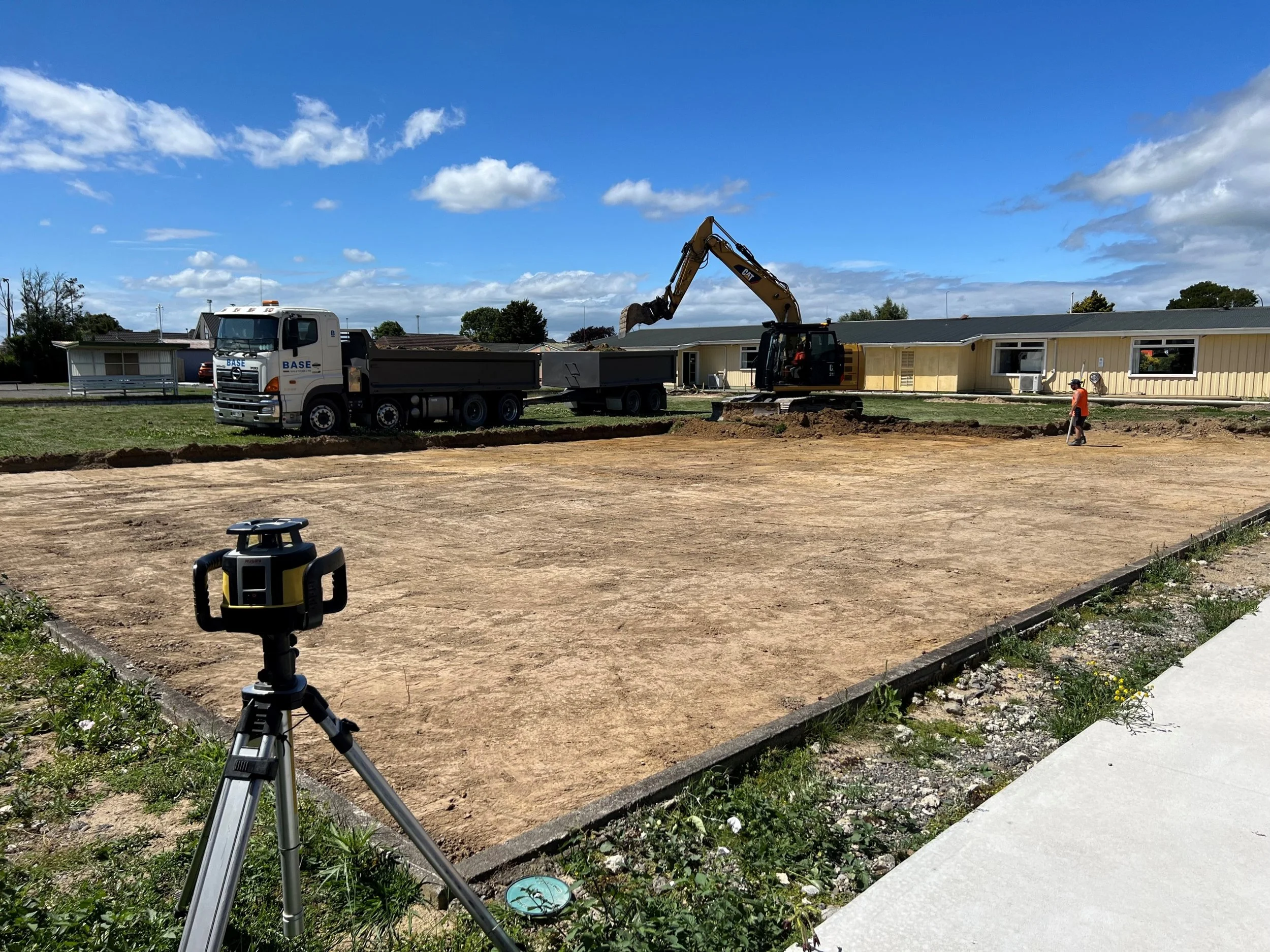 Construction site with bulldozer and dump truck on cleared dirt lot, a worker in orange vest, survey equipment on tripod, and residential buildings in the background under a partly cloudy sky.
