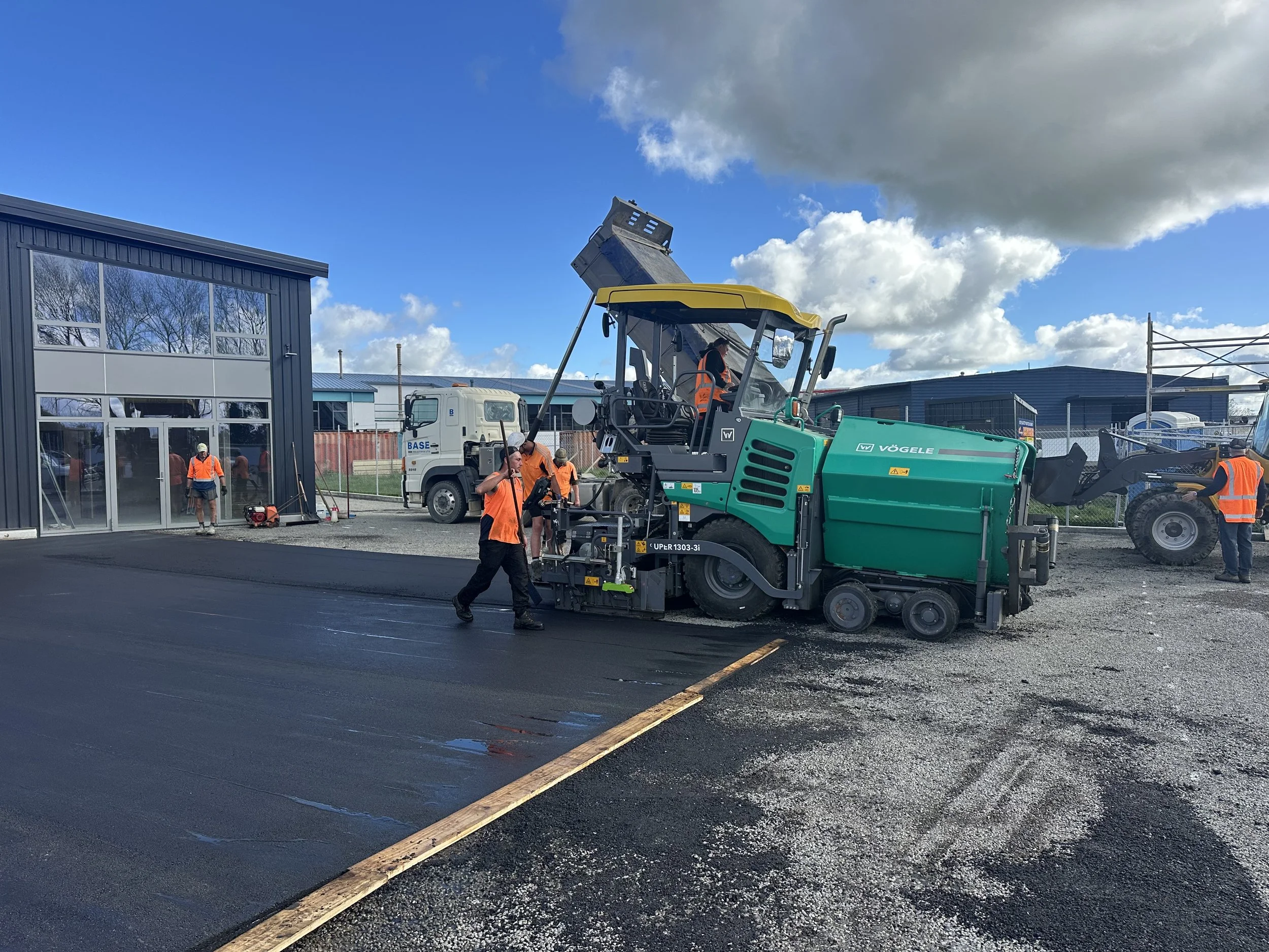 Workers in orange safety vests paving a road with asphalt; some workers are operating machinery while others are walking or observing, with a building and construction site in the background under a partly cloudy sky.