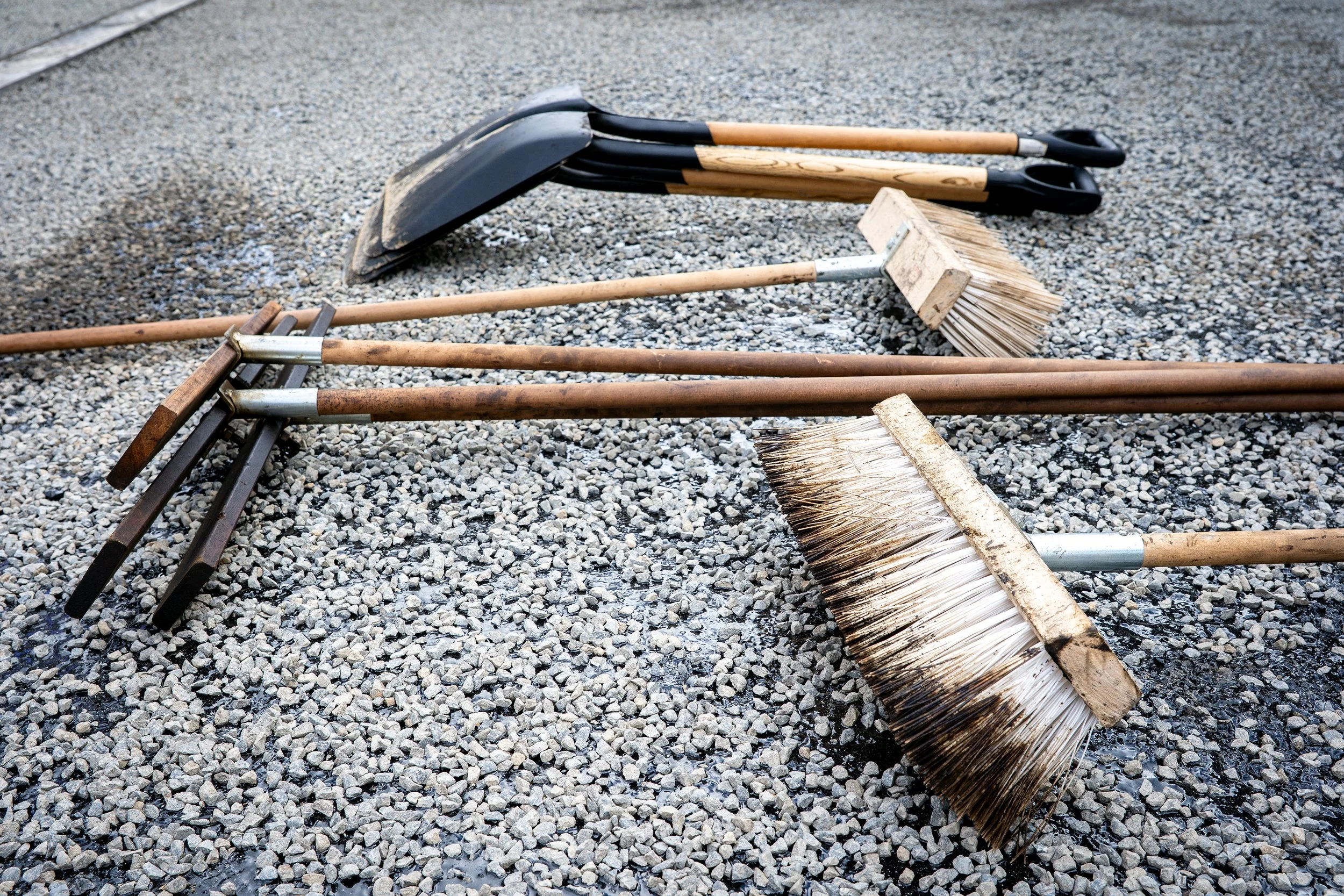 A collection of outdoor cleaning tools, including shovels, brooms, and a rake, lying on gravel ground.