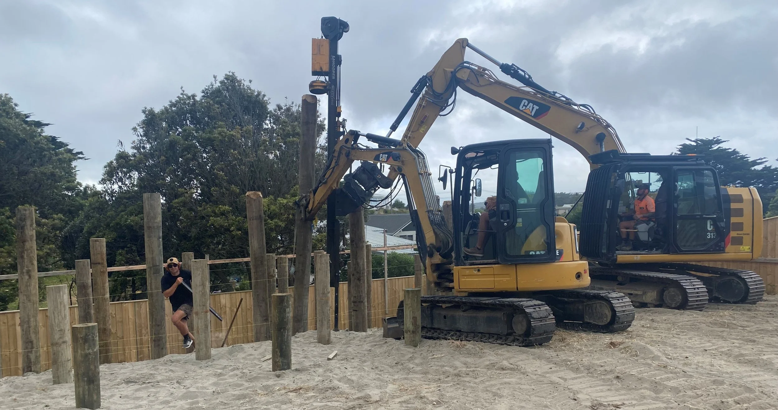 Construction site with two yellow excavators working on wooden posts, cloudy sky, person in sunglasses and shorts near posts, residential neighborhood in background.