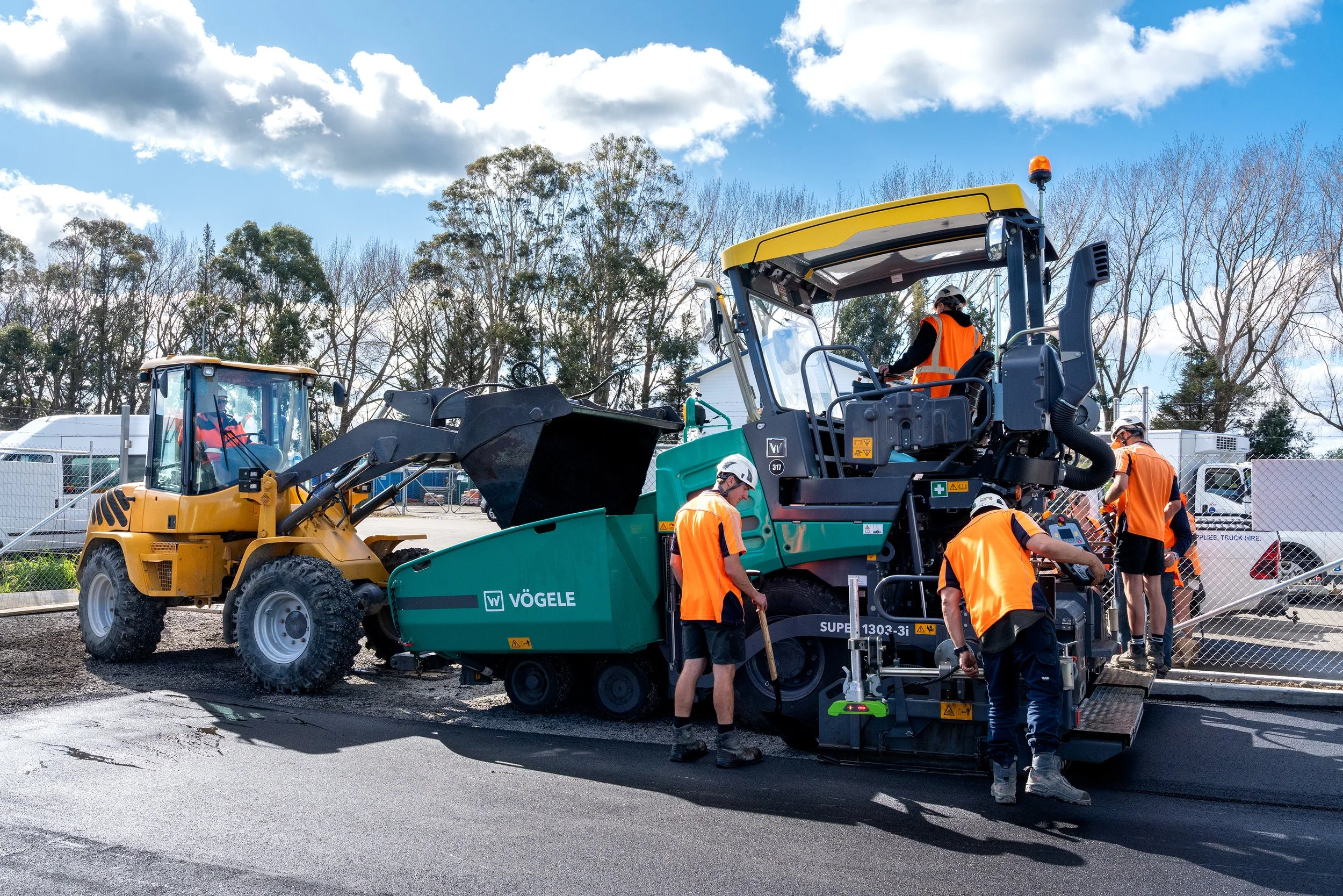 Construction workers in orange safety vests and helmets operating paving machinery on a road under a partly cloudy sky.