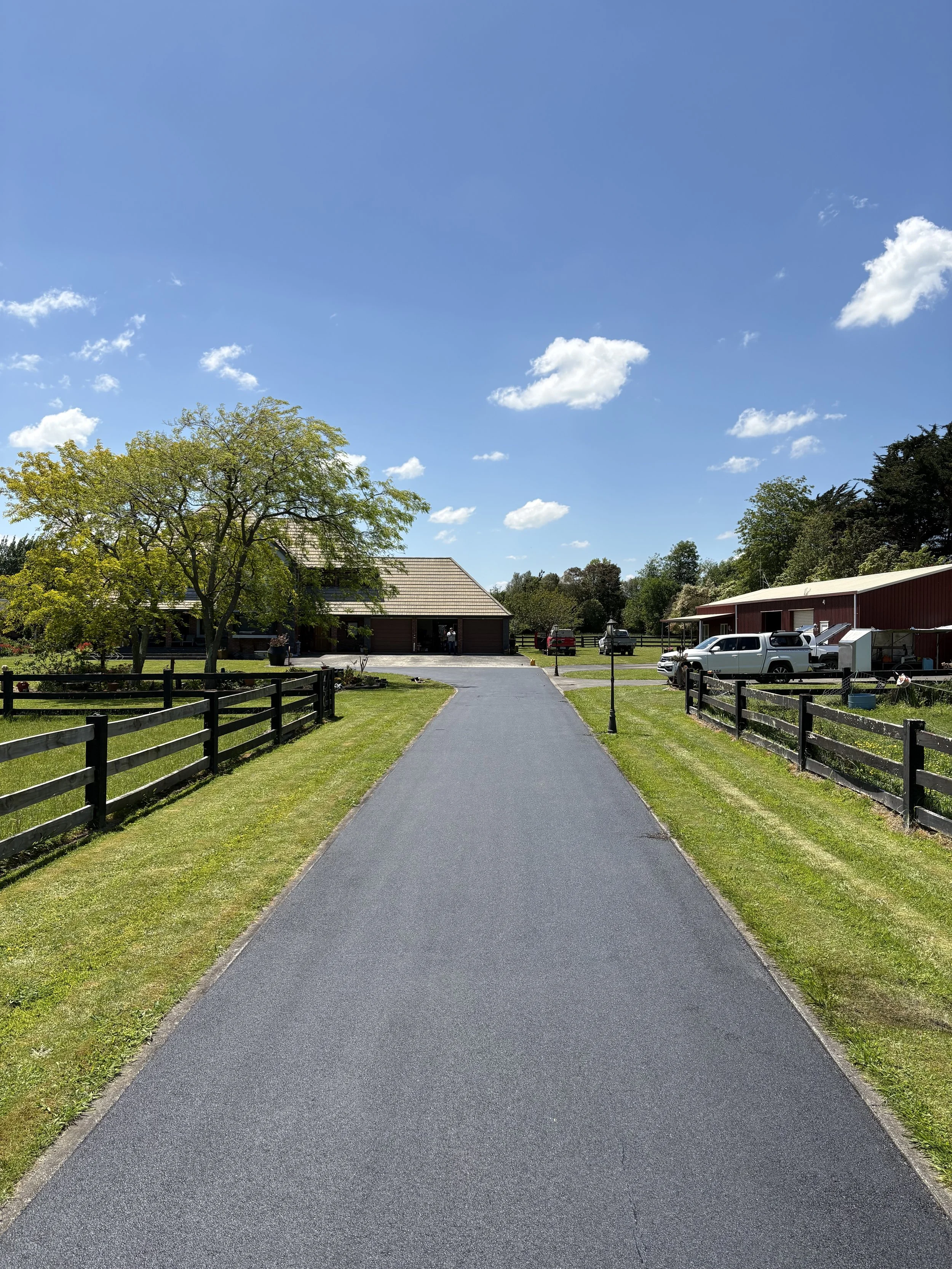A paved driveway leading to a house, with a grassy lawn and black wooden fences on both sides, a tree with green leaves on the left, parked vehicles on the right, and a blue sky with scattered clouds.