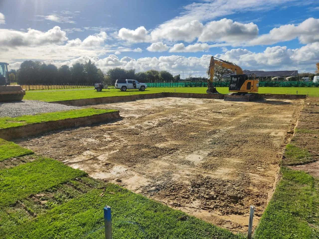 Construction site with excavator working on foundations, surrounded by parked vehicles, green grass, and a blue sky with clouds.