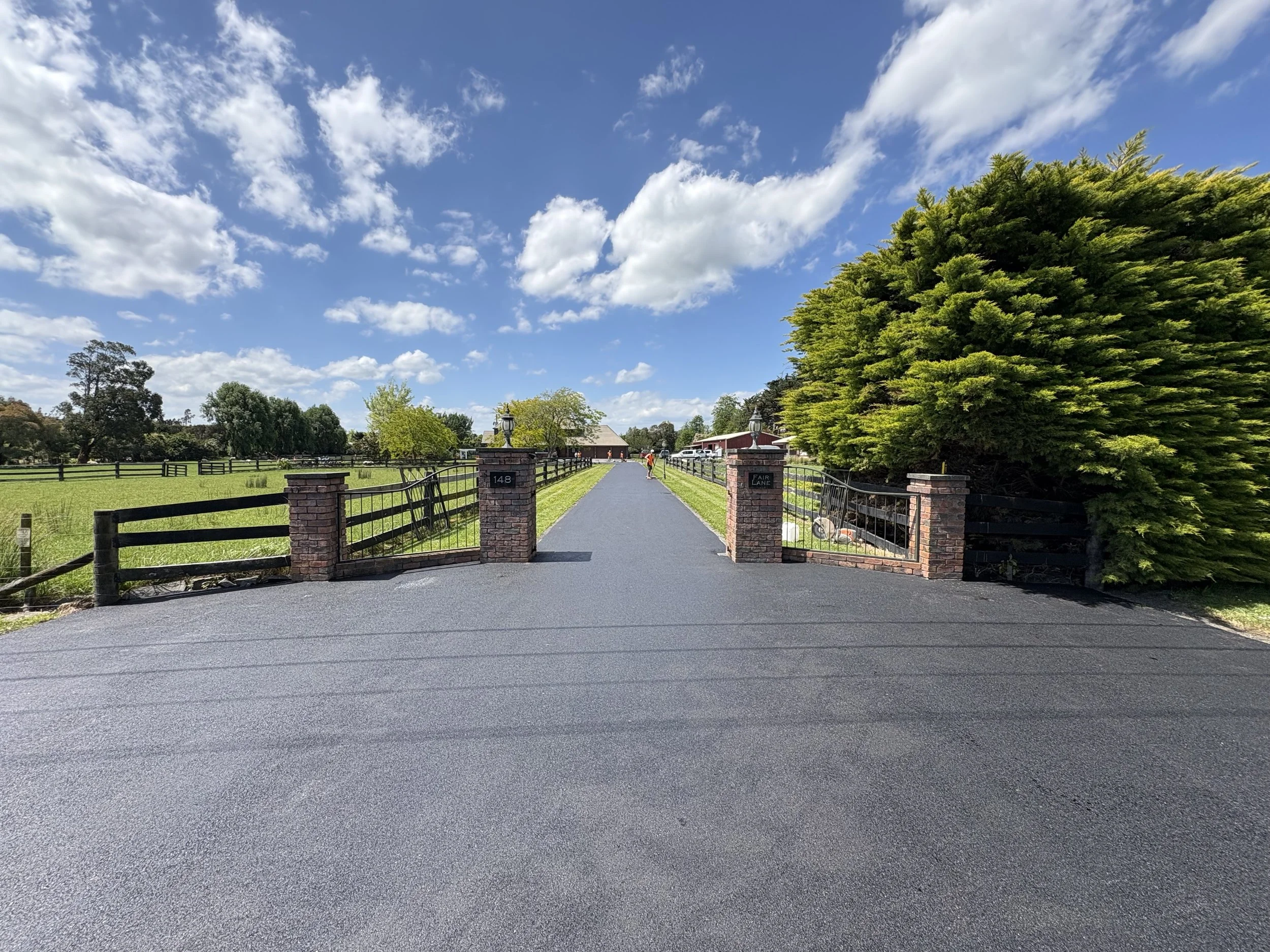 A paved driveway with brick pillars and metal gates leading to a property, with green trees and a blue sky with fluffy clouds in the background.