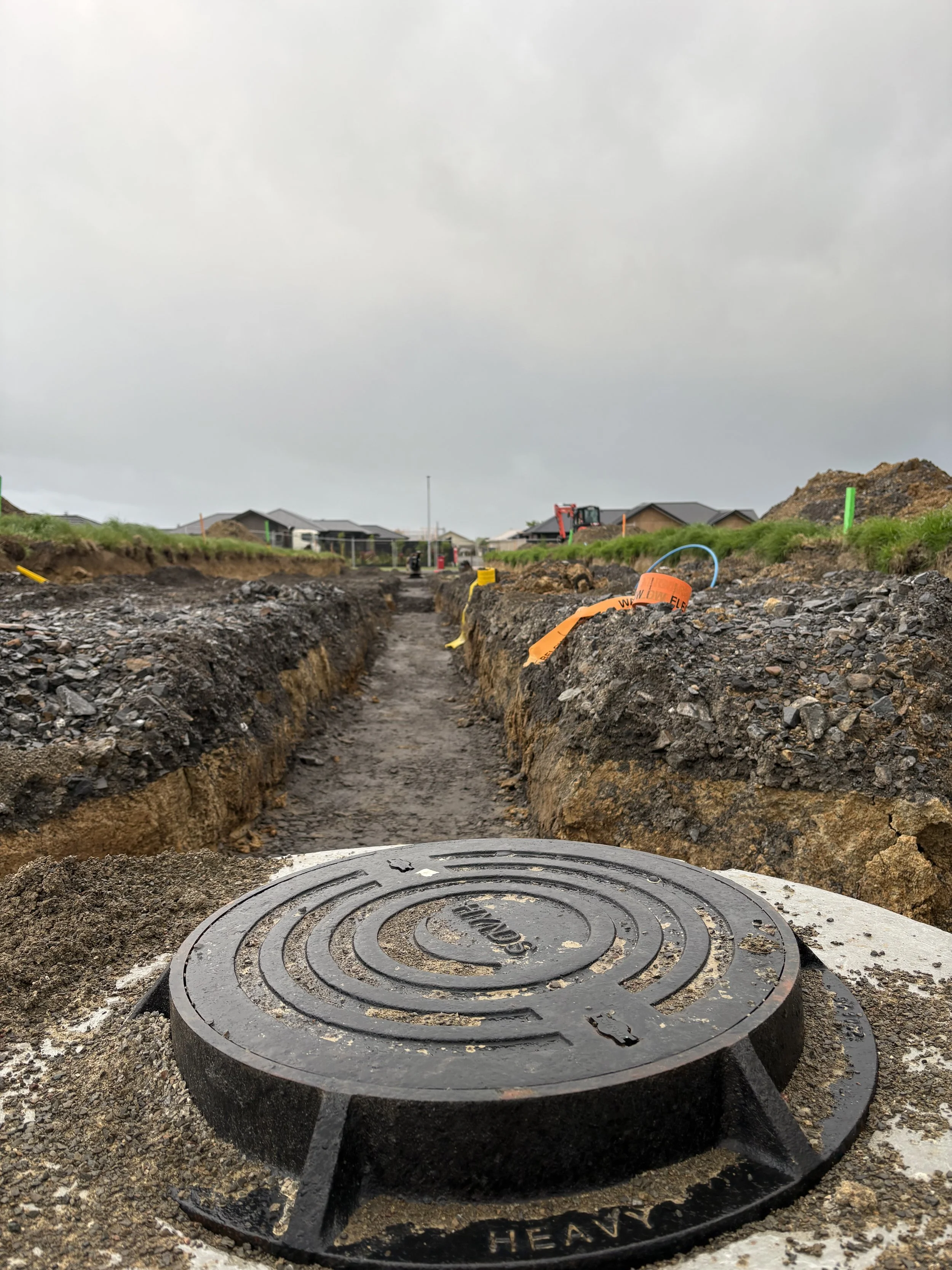 View of a construction site with a dug trench, a heavy manhole cover in the foreground, and construction equipment and houses in the background under an overcast sky.
