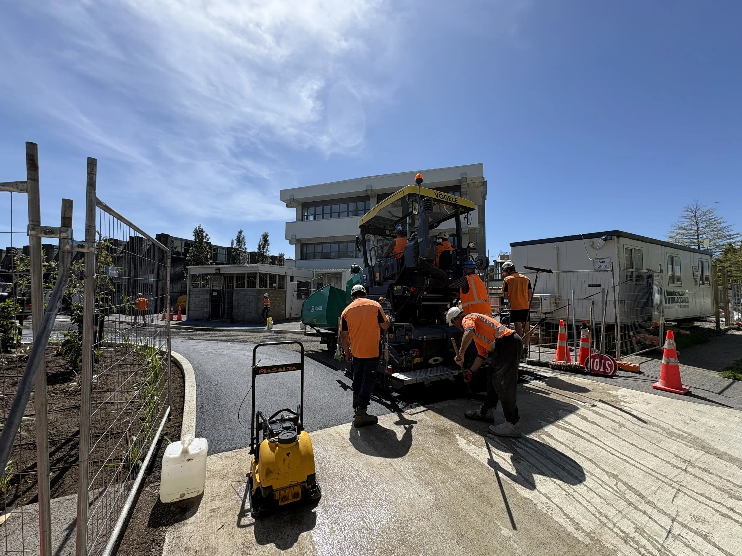 Construction workers in orange safety vests and helmets operating machinery and working on a road near a construction site with orange cones and fencing.