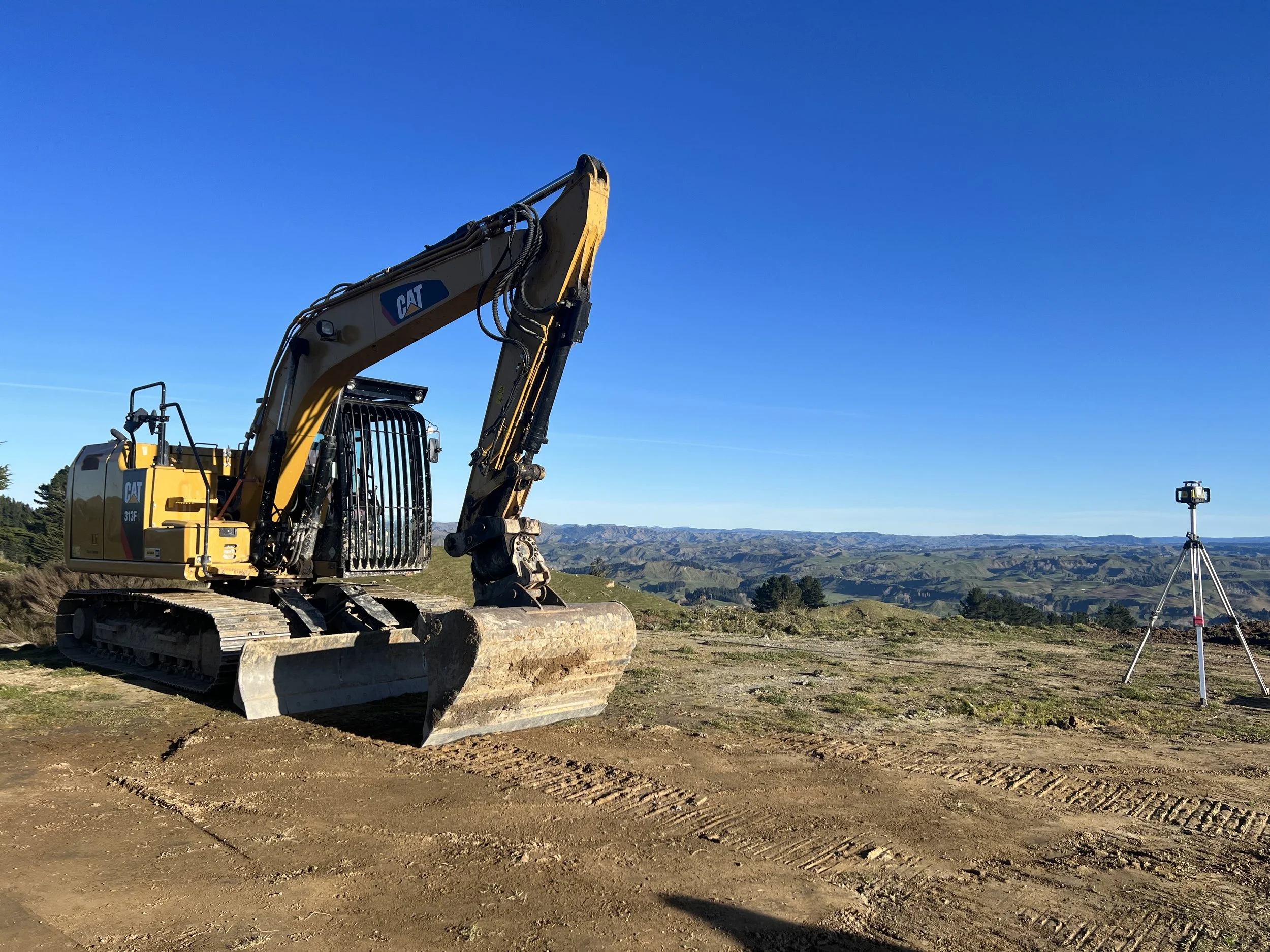 A yellow excavator is parked on a dirt construction site with hills in the background. A surveying instrument on a tripod is positioned to the right of the excavator under a clear blue sky.