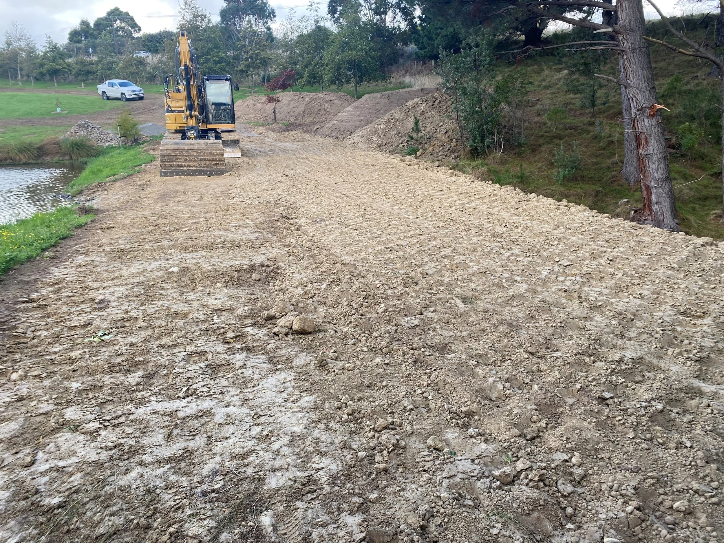 A construction site with a yellow bulldozer working on a gravel pathway near a small pond, with trees and grassy areas in the background.