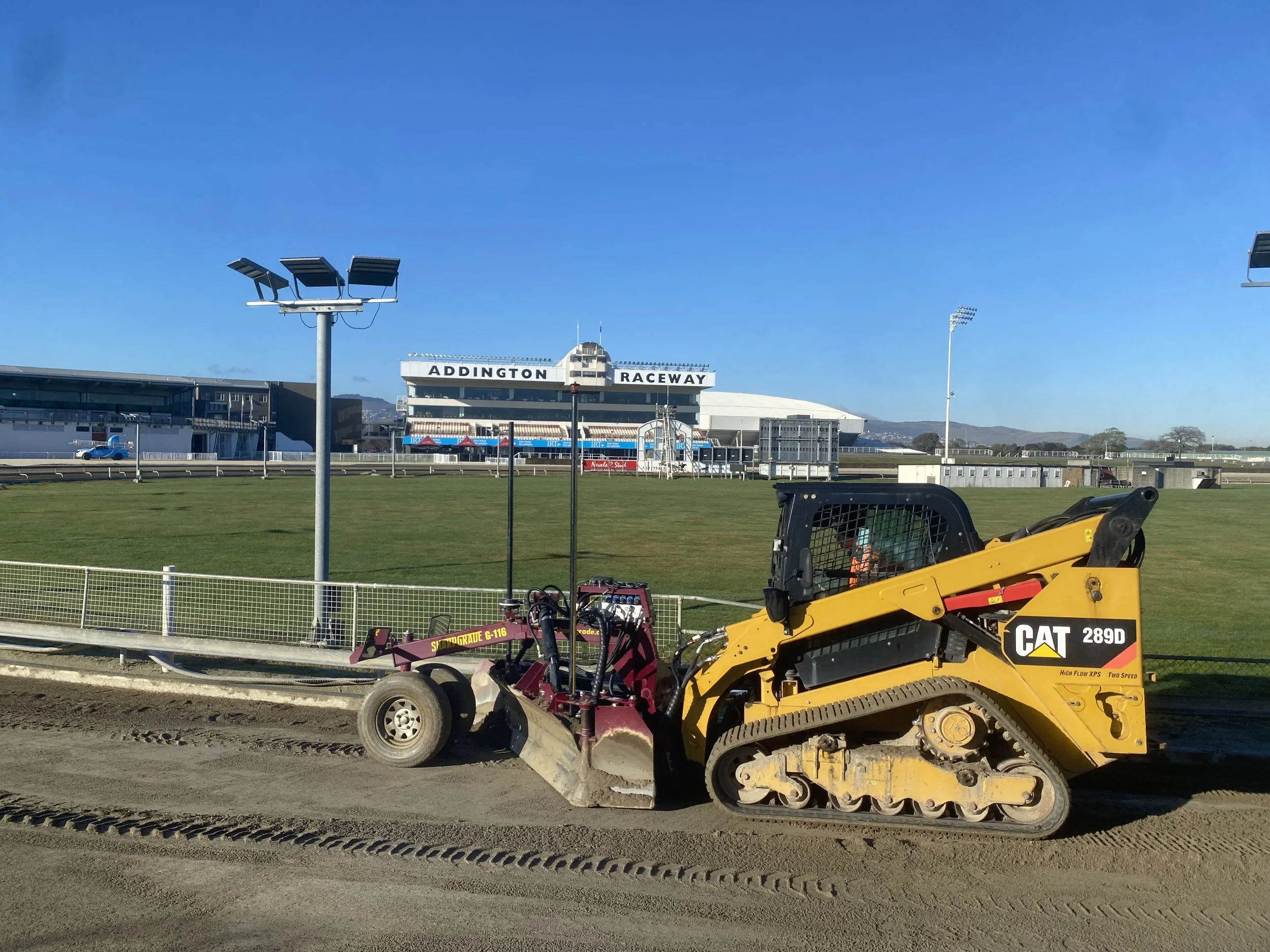 A construction vehicle, a yellow Caterpillar 289D compact track loader, working on the dirt track at the racetrack with the building labeled "Addington Raceway" in the background.
