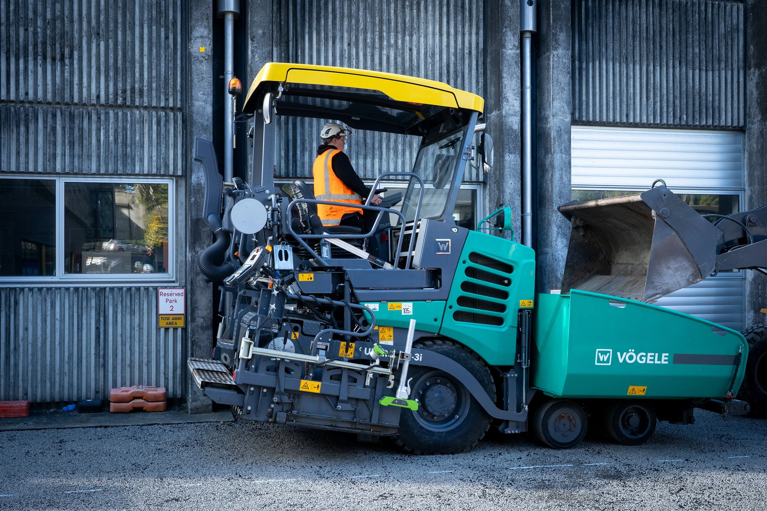 A construction worker operating a teal and yellow street sweeper machine in front of a gray industrial building with windows.