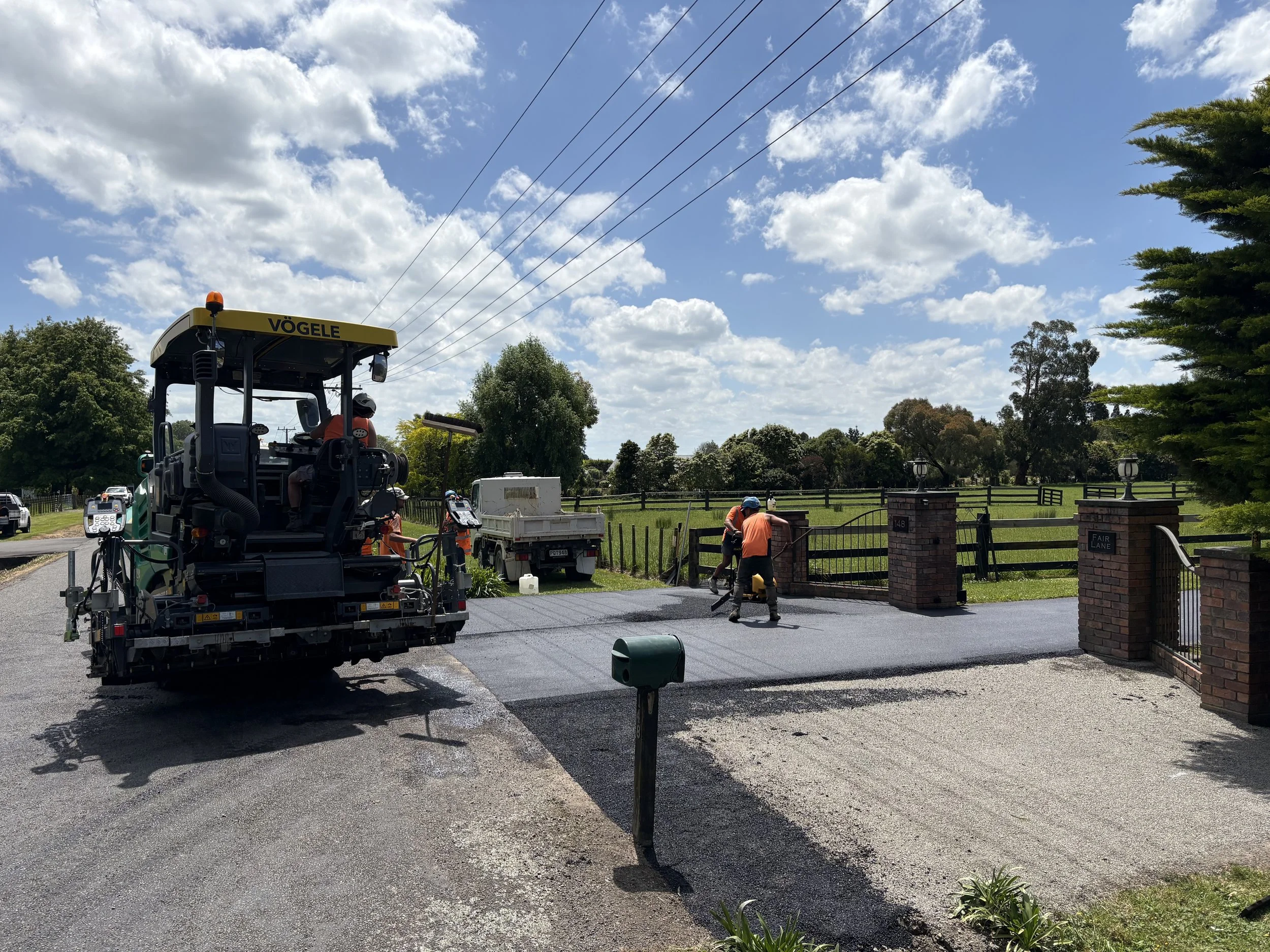 Workers paving a driveway in front of a brick entrance gate, with a pickup truck and construction equipment under a partly cloudy sky.