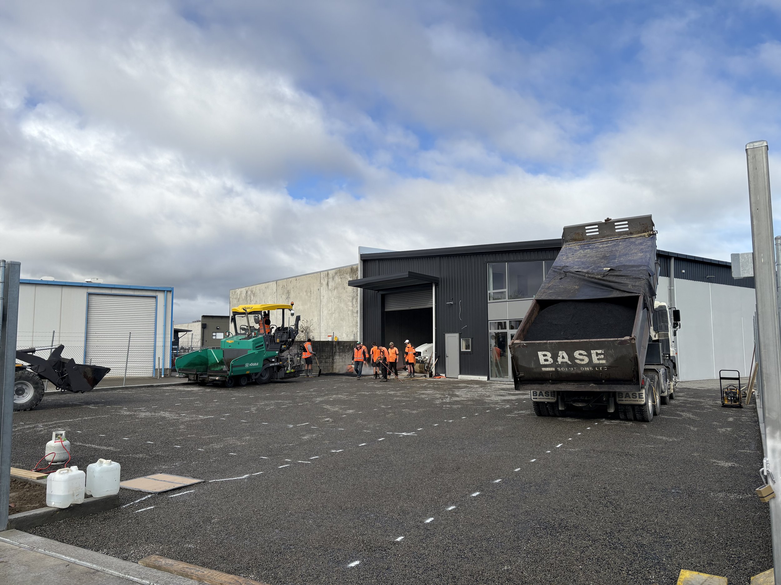 Construction workers in orange vests working outside a building with construction vehicles, including a dump truck and a road roller, on a gravel lot with marked parking spaces, under a partly cloudy sky.