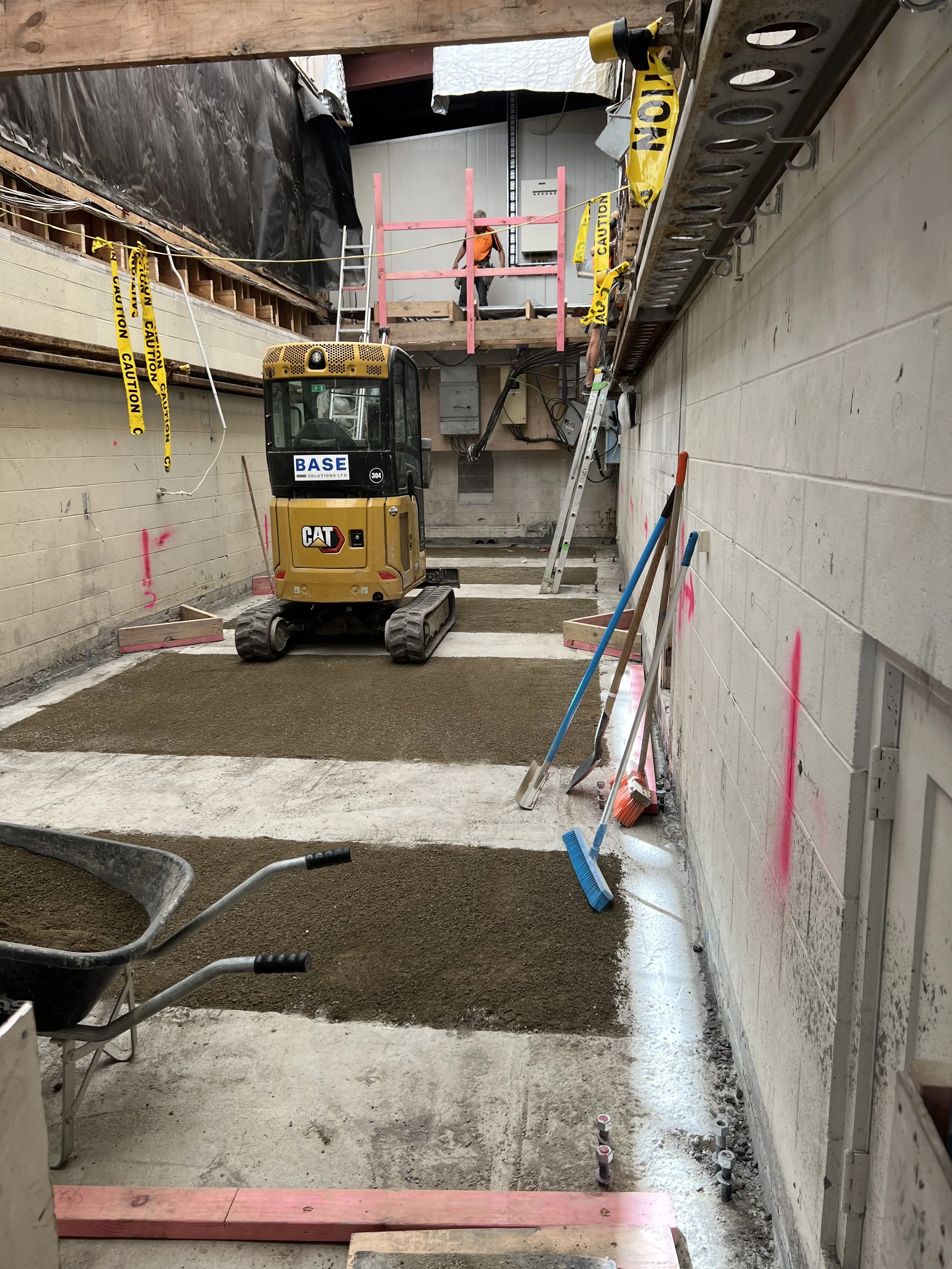 Partially constructed interior space with construction equipment and tools, including a small yellow CAT excavator, a wheelbarrow, shovels, and brooms, with pink markings on the walls and safety caution tape hanging nearby.