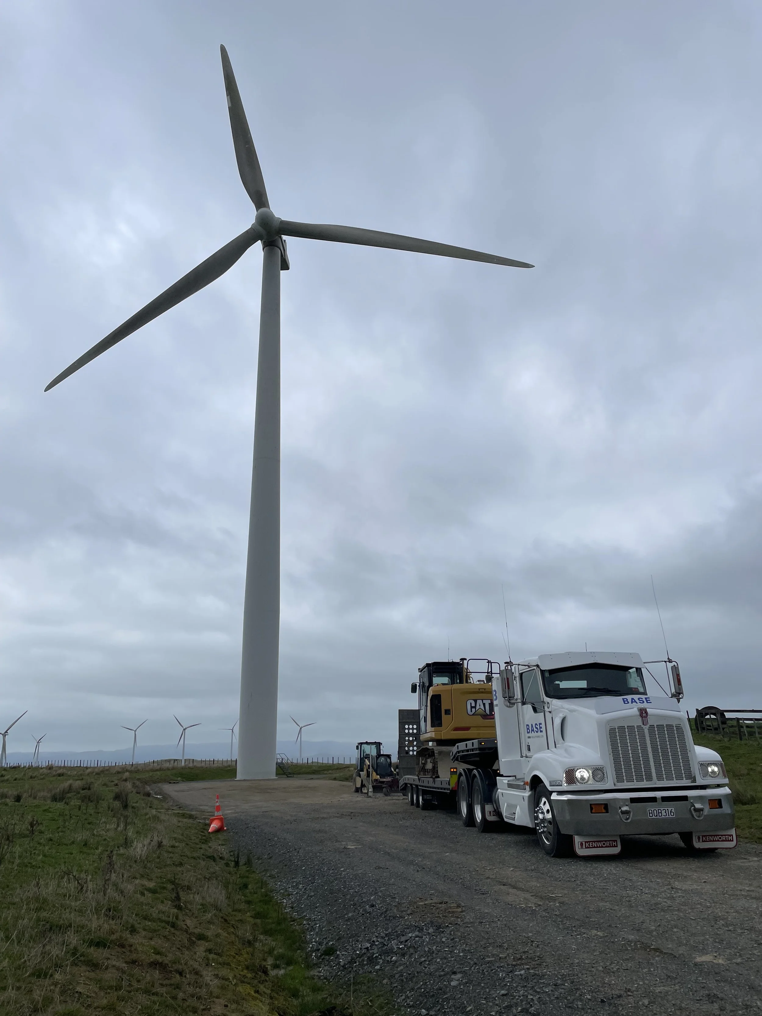 A large wind turbine under construction with a truck and construction equipment nearby on a gravel road, under a cloudy sky.