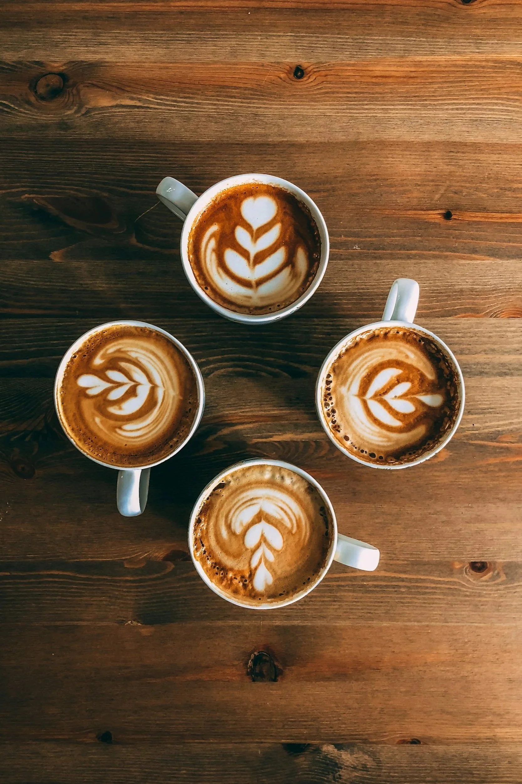 Four cups of coffee with latte art on a wooden table, viewed from above.