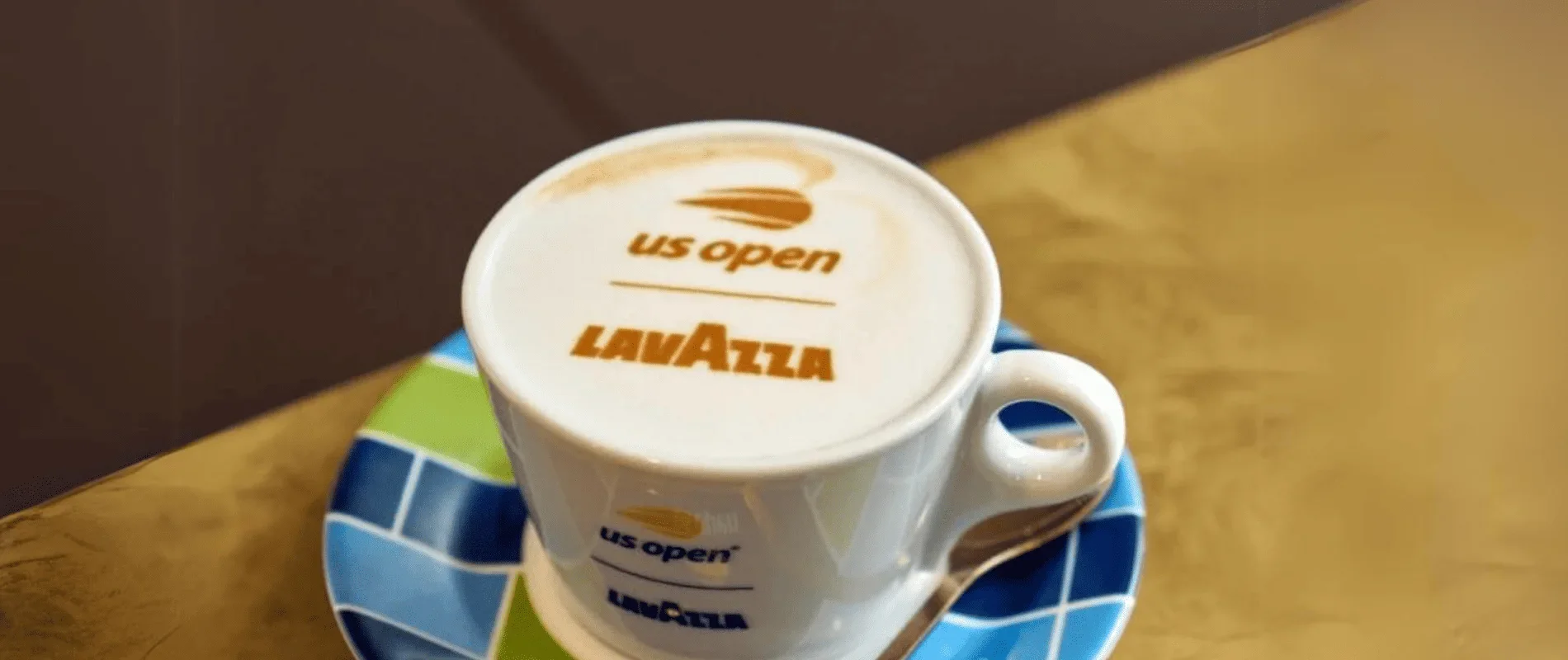 Coffee cup with foam art displaying the words 'us open' and 'LAVAZZA' on top, sitting on a colorful blue and green saucer on a wooden table.