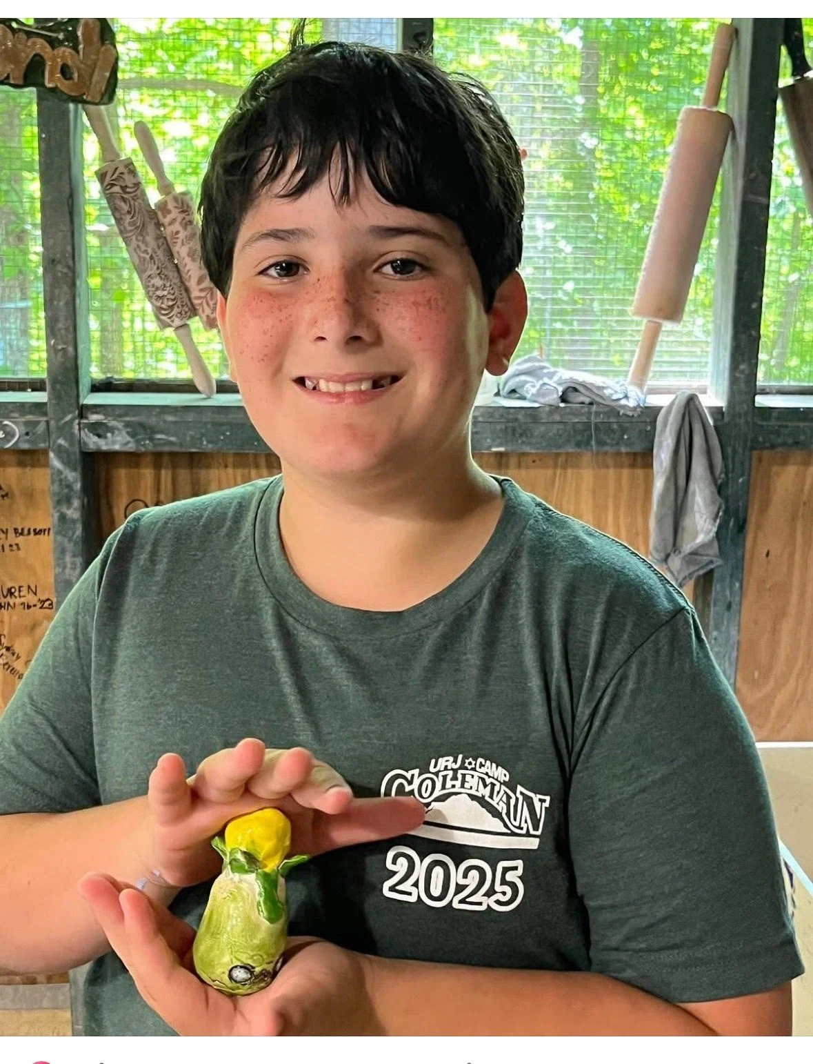 A boy with dark hair and freckles smiling at the camera, holding a small object wrapped in yellow and green paper, inside a rustic wooden shed with rolling pins hanging on the wall behind him.