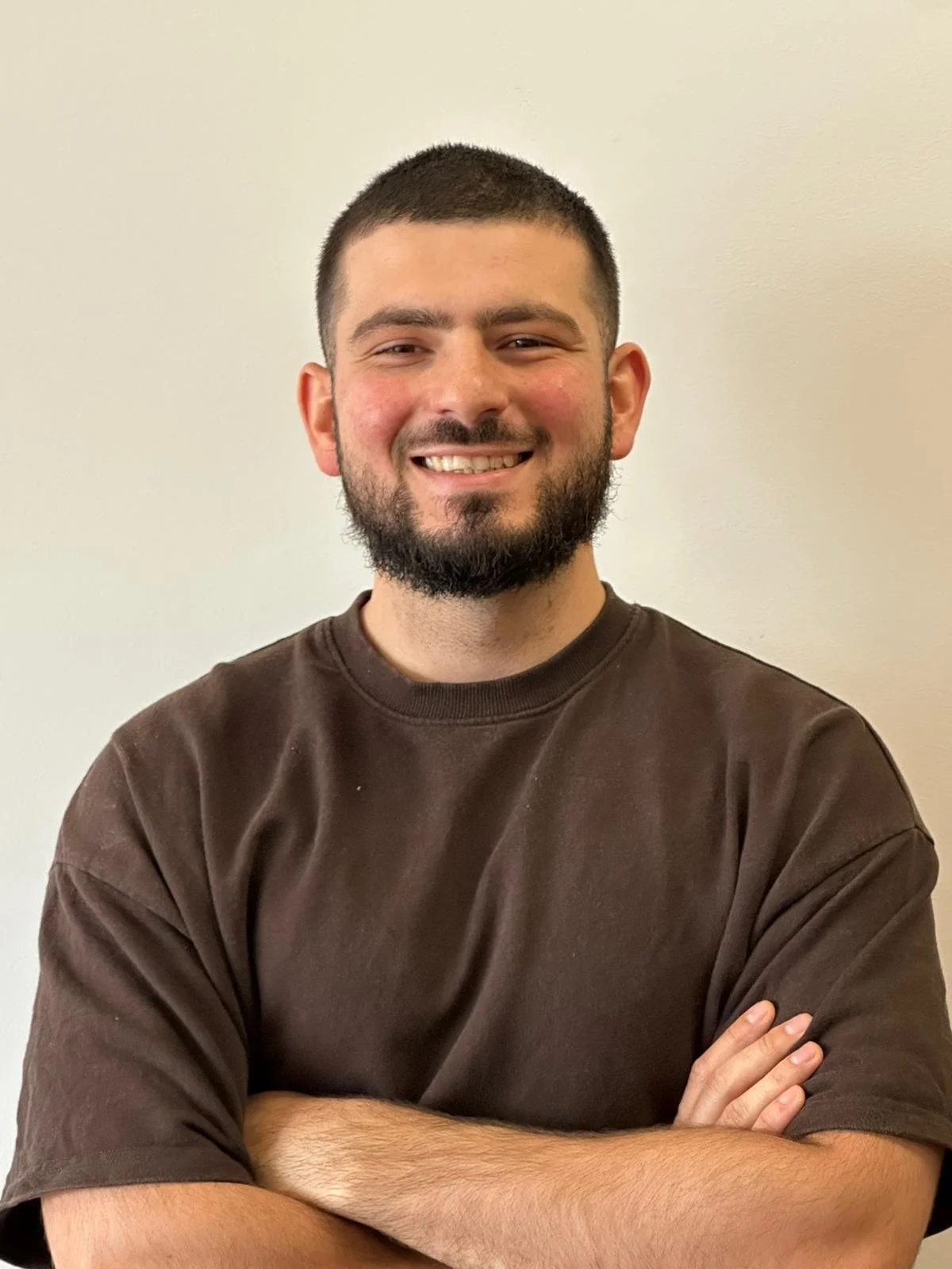 A man with curly dark hair, glasses, a nose ring, and a beard, wearing a dark green sweater.
