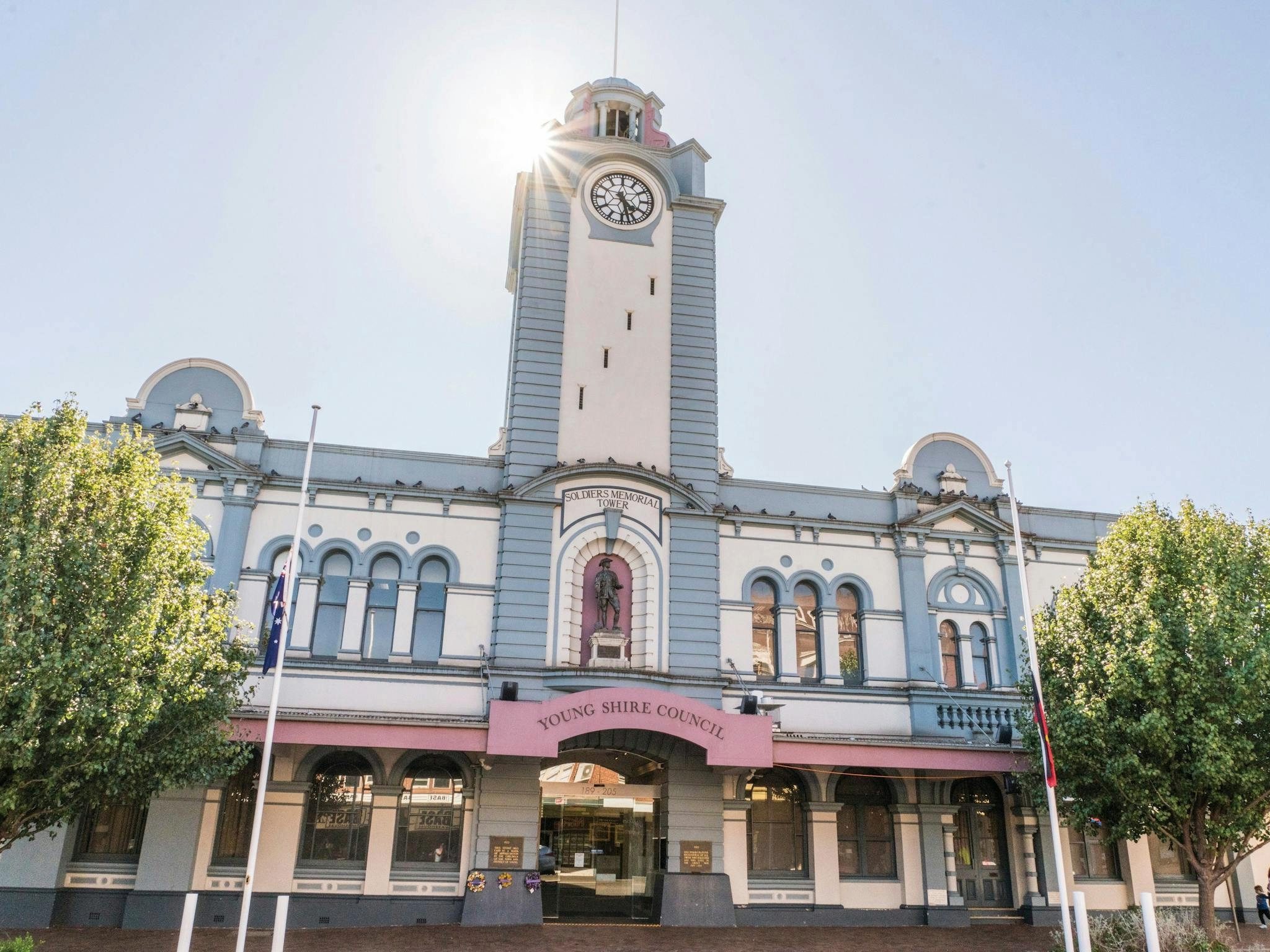 Front view of the Soldiers Memorial Tower building with a pink sign that reads 'Young Shire Council', trees on either side, and bright sunlight creating lens flare.