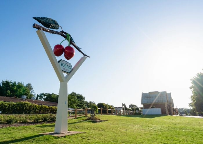 Large vertical sign in a park with a giant fork, cherries, a grass field, and a wooden fence, with buildings and trees in the background under a clear blue sky.