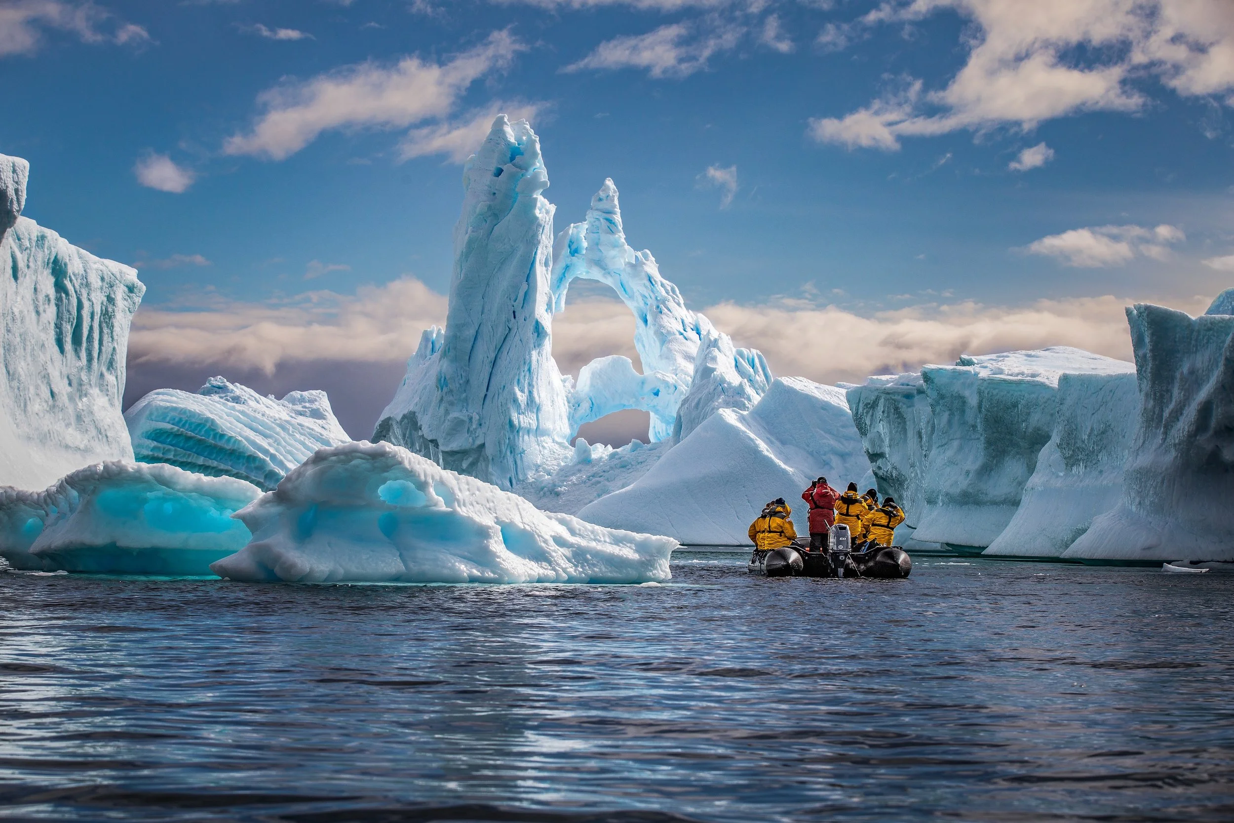 A group of people in yellow and red jackets on a boat near large icebergs and ice formations in icy waters under a partly cloudy sky.