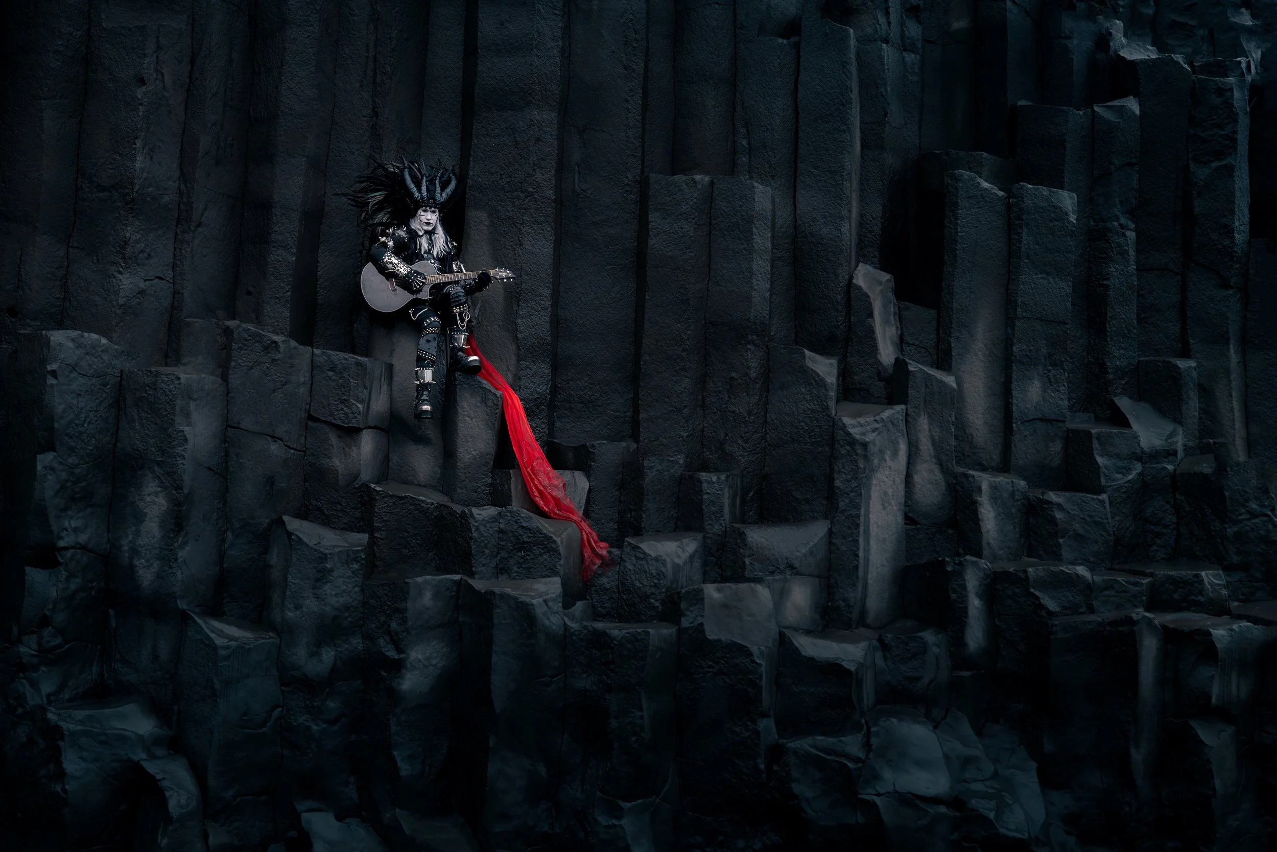 A person dressed in gothic style with black spiked hair, black leather outfit, and face paint, sitting on dark rocks while playing an acoustic guitar. There is a long red cloth hanging down from the rocks.