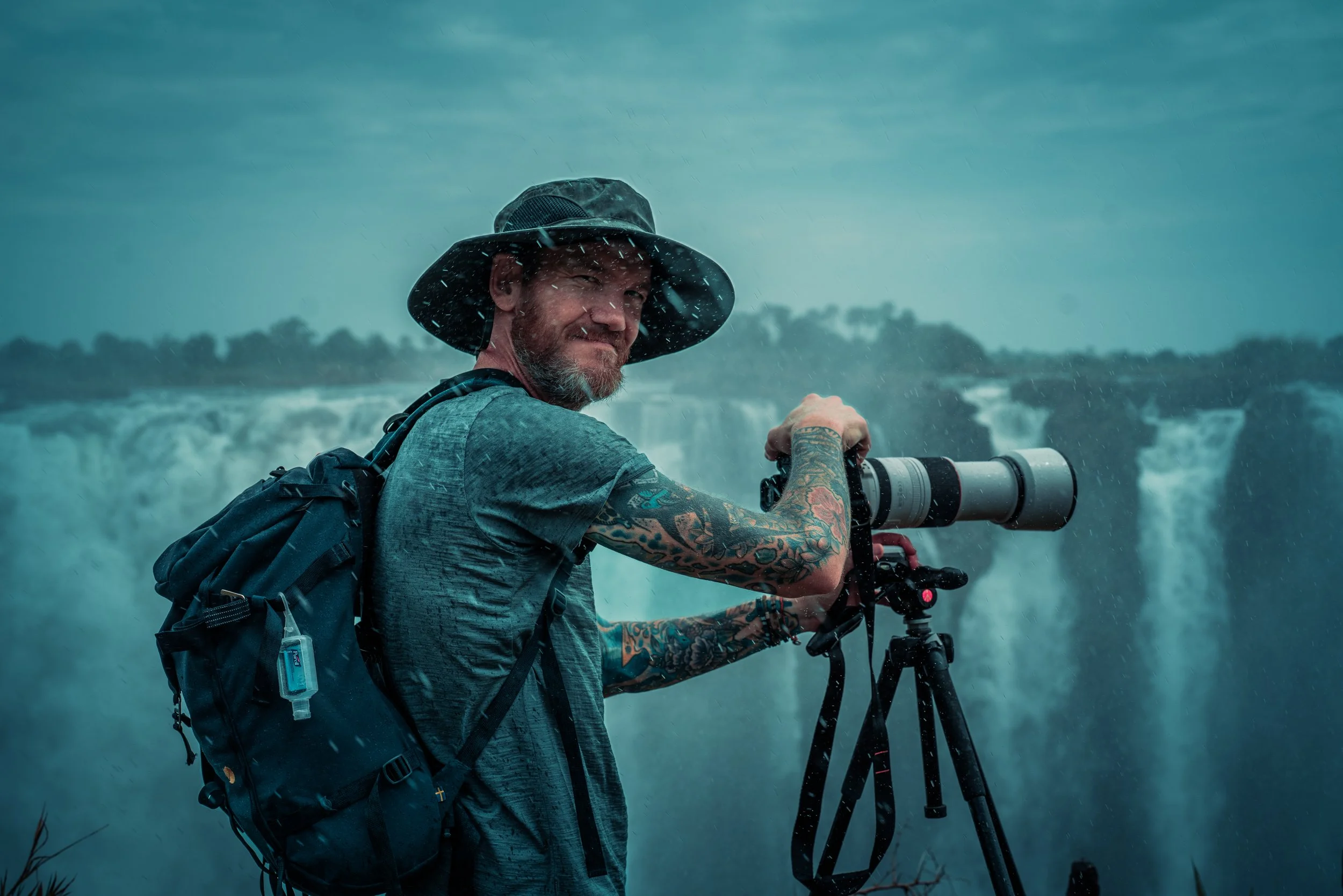 Photographer standing outdoors near waterfalls, holding a camera with a long lens, wearing a wide-brimmed hat, a backpack, and a gray shirt, with rain and clouds overhead.
