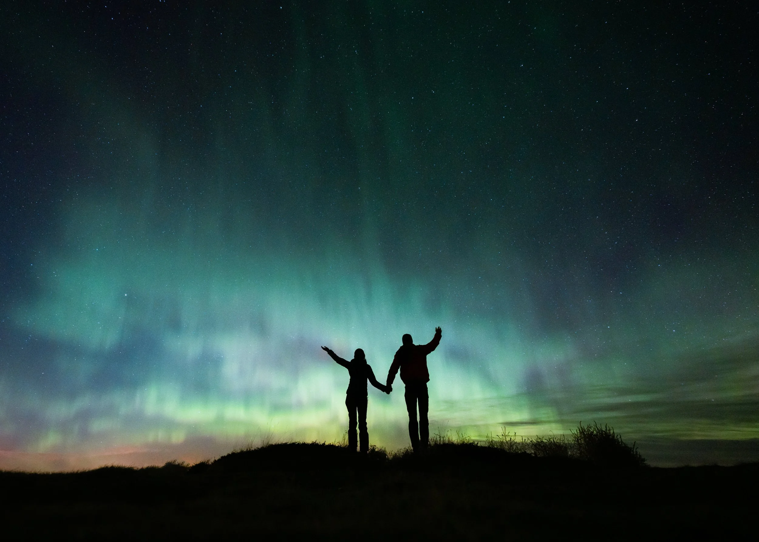 Silhouettes of two people holding hands and raising their arms in front of a vibrant green and blue Northern Lights display in a starry night sky.