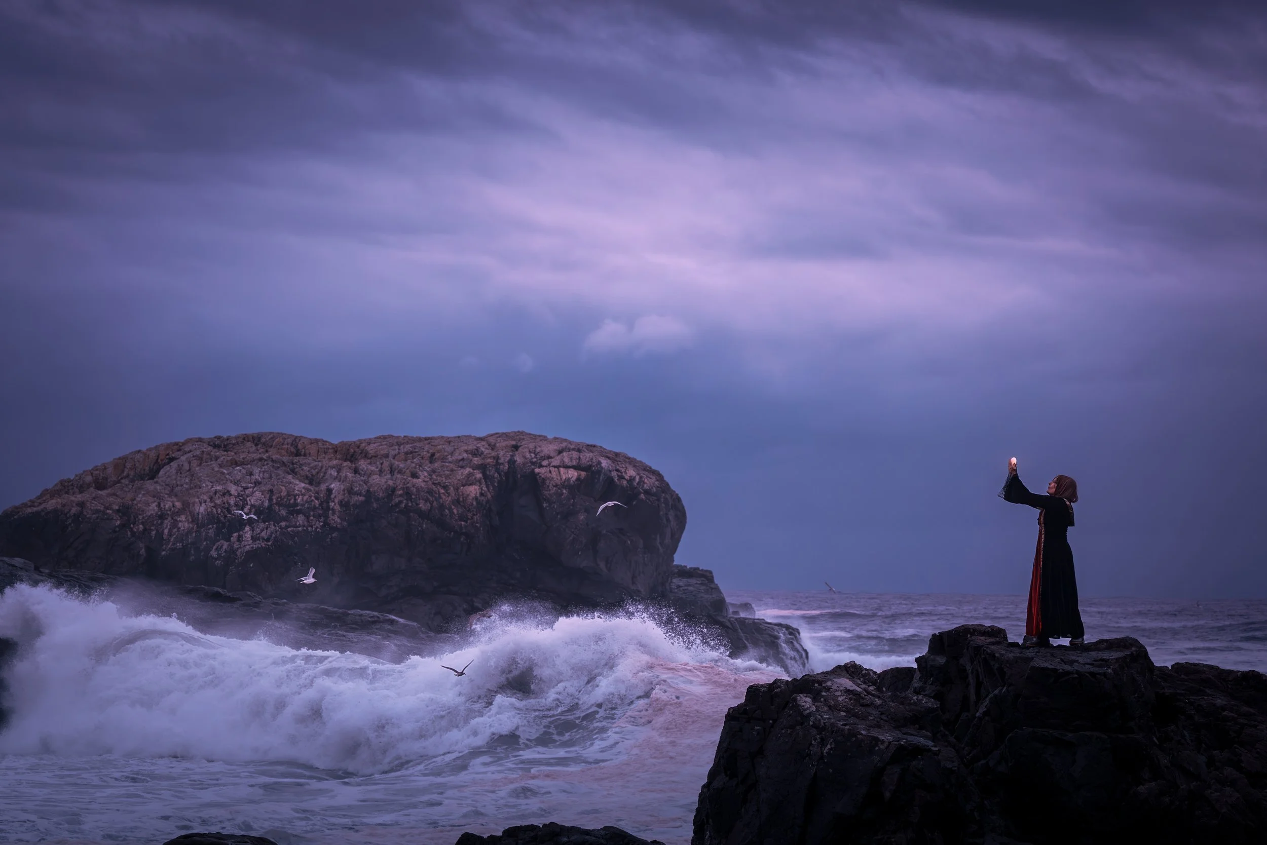 A woman standing on rocks at the ocean, taking a photo with her phone as waves crash against the rocks and seagulls fly overhead under a cloudy, purple-toned sky.