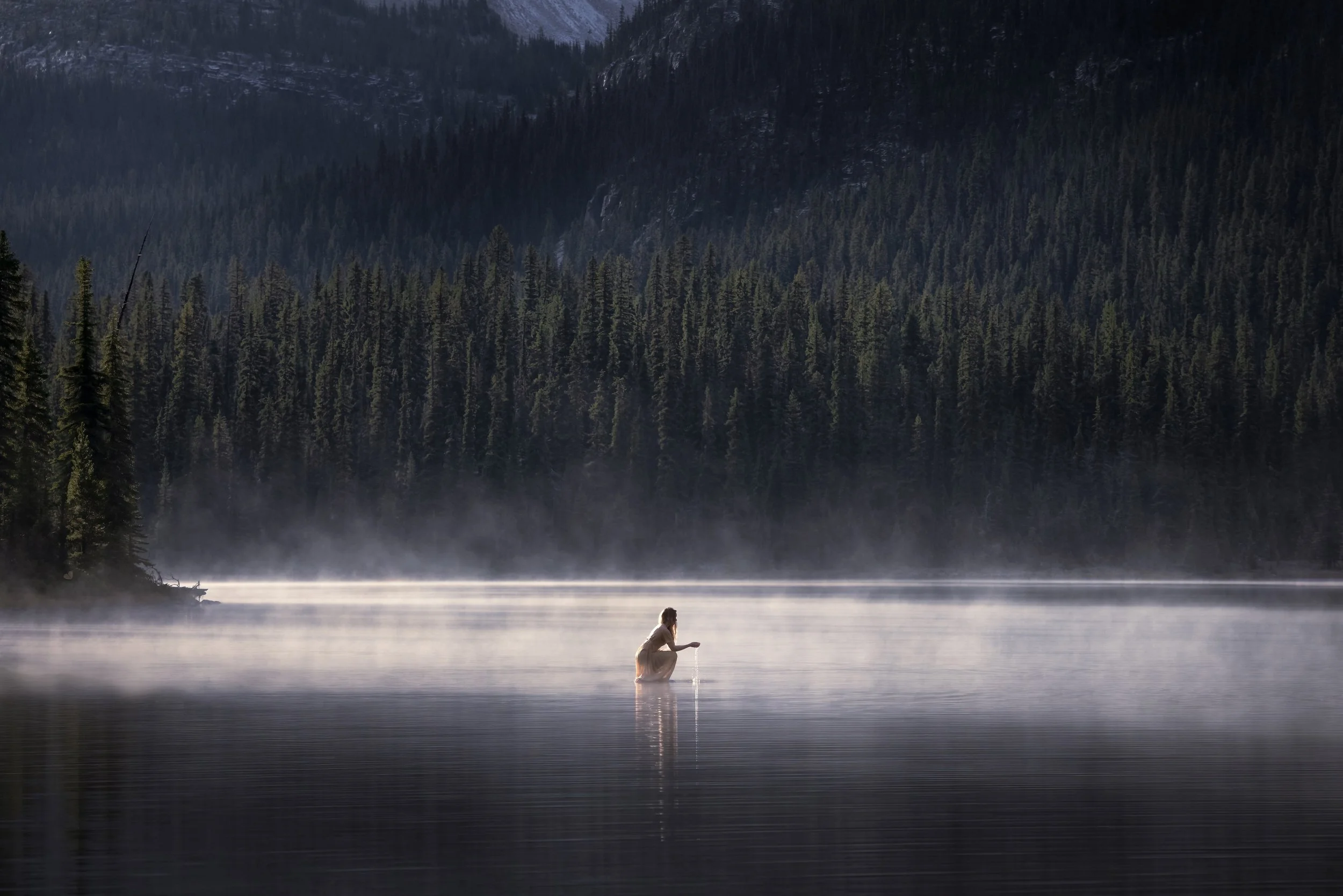 Person standing in a calm lake with mist, surrounded by a dense forest and mountains in the background.