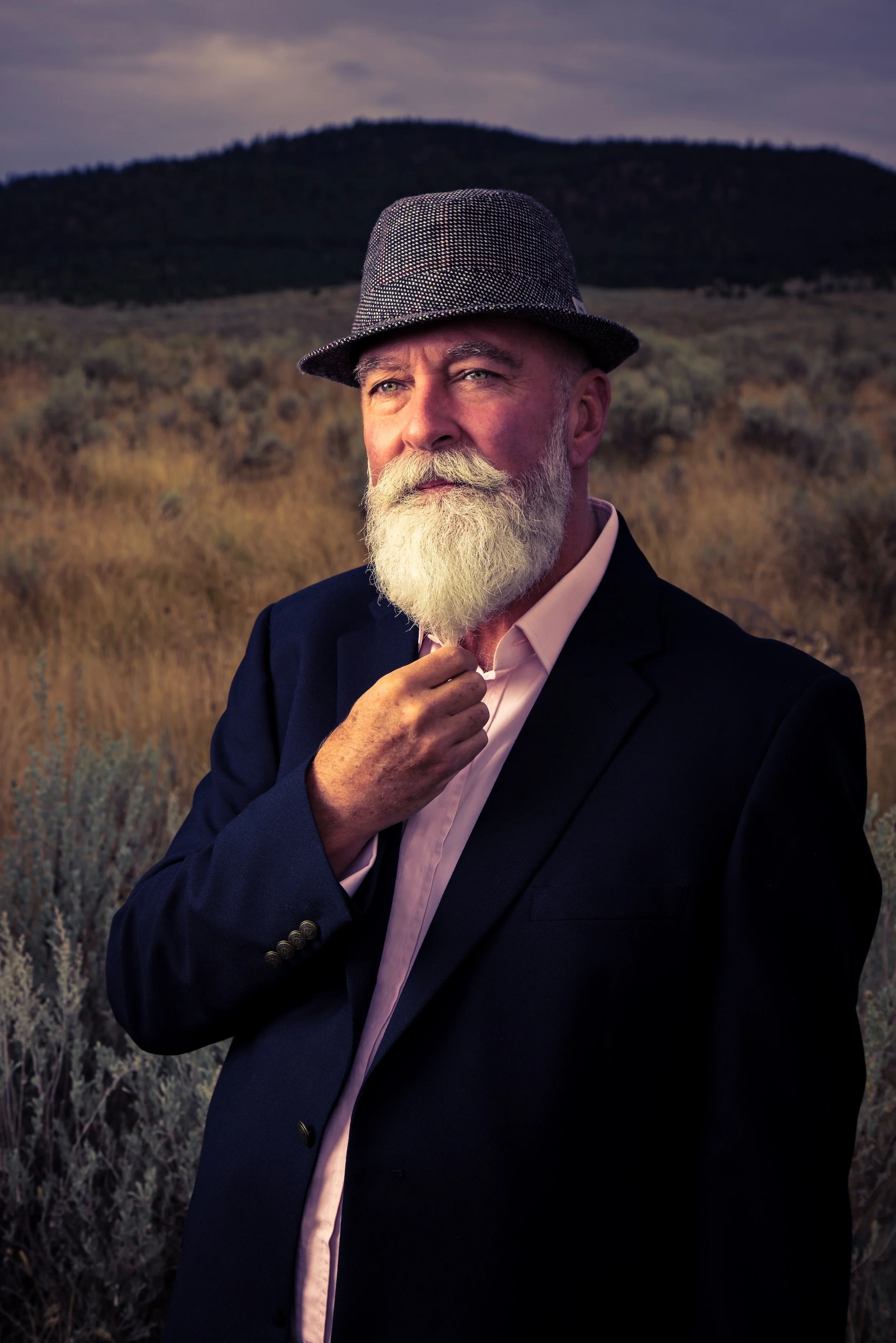 A man with a white beard and mustache, wearing a black suit jacket, a light pink shirt, and a gray fedora hat, standing outdoors in a field with mountains in the background, during sunset.