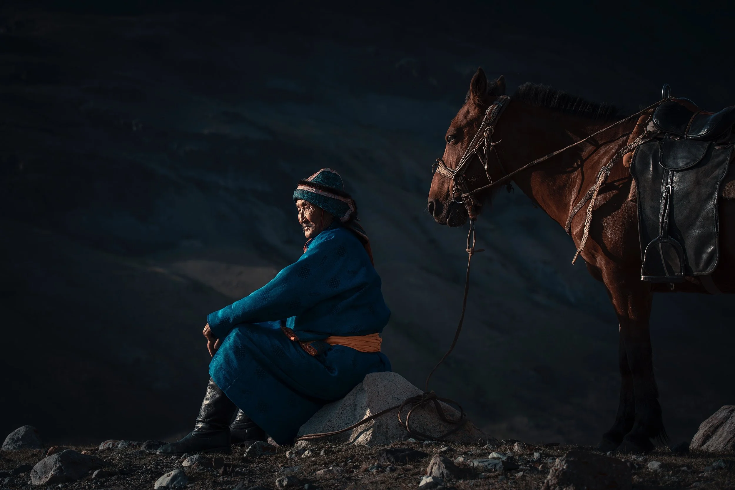An elderly man dressed in traditional Mongolian clothing sitting on a rock next to a brown horse, with a dark mountain landscape in the background.