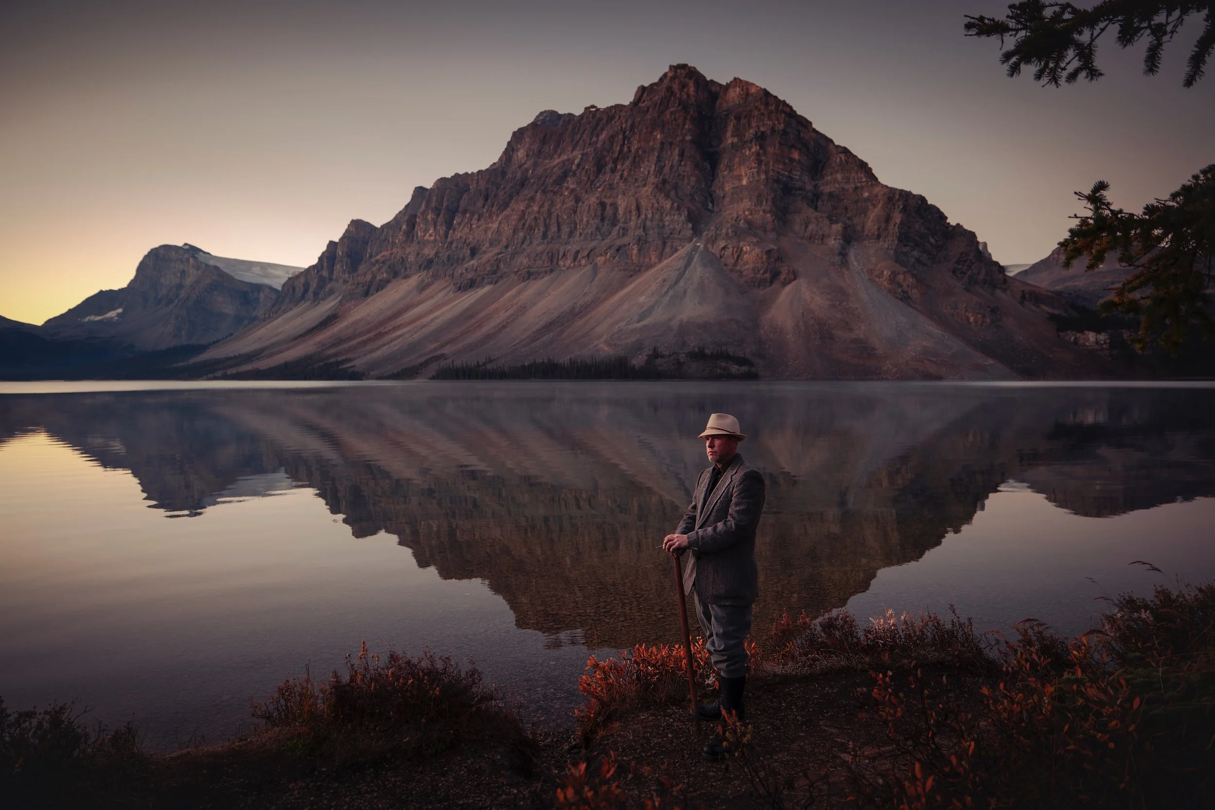 A man in vintage clothing, wearing a hat, stands by a calm lake surrounded by mountains at dawn or dusk, with a reflection of the mountains in the water.