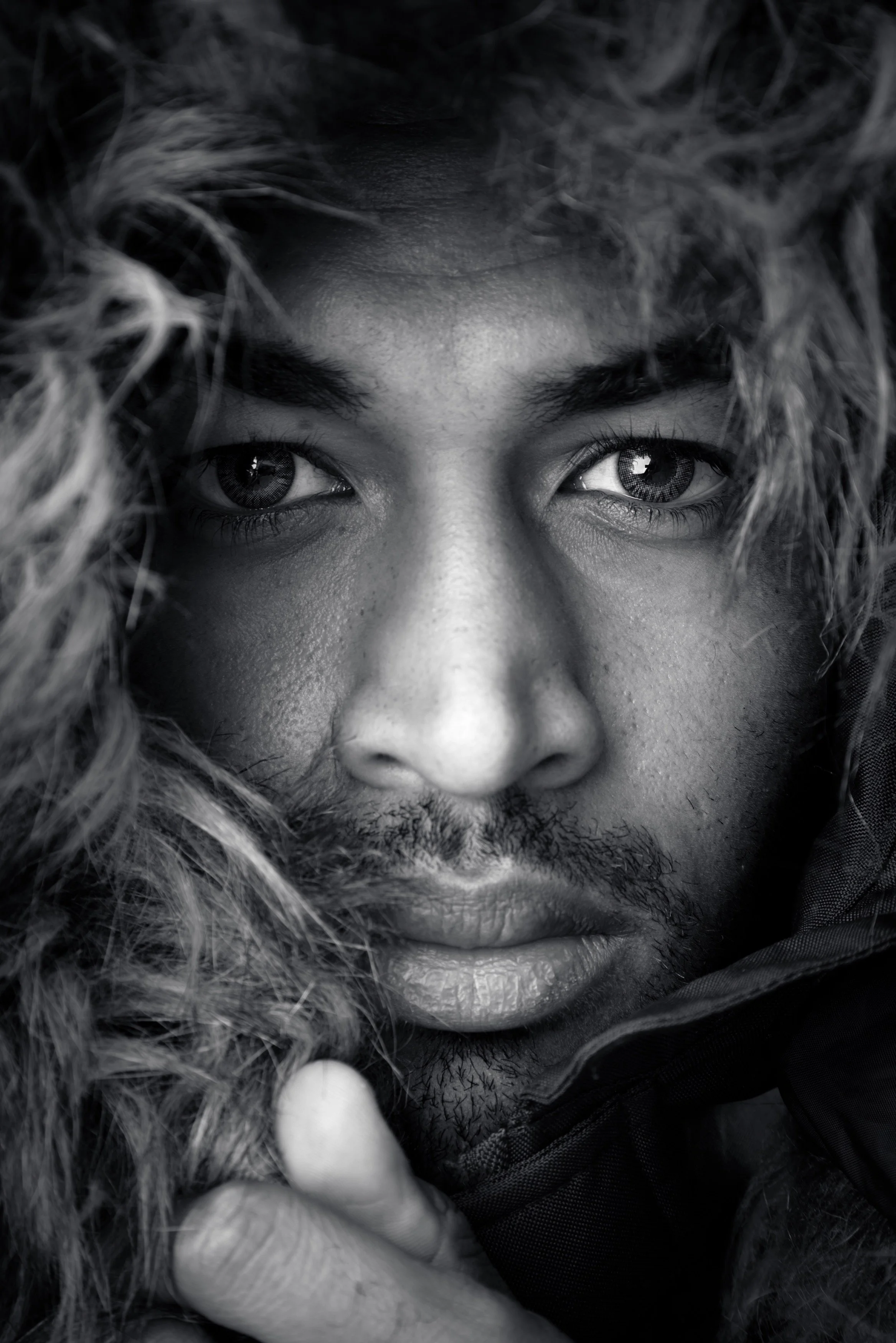 Close-up black and white photo of a person's face with light-colored eyes, curly hair, and full lips, wearing a dark jacket.