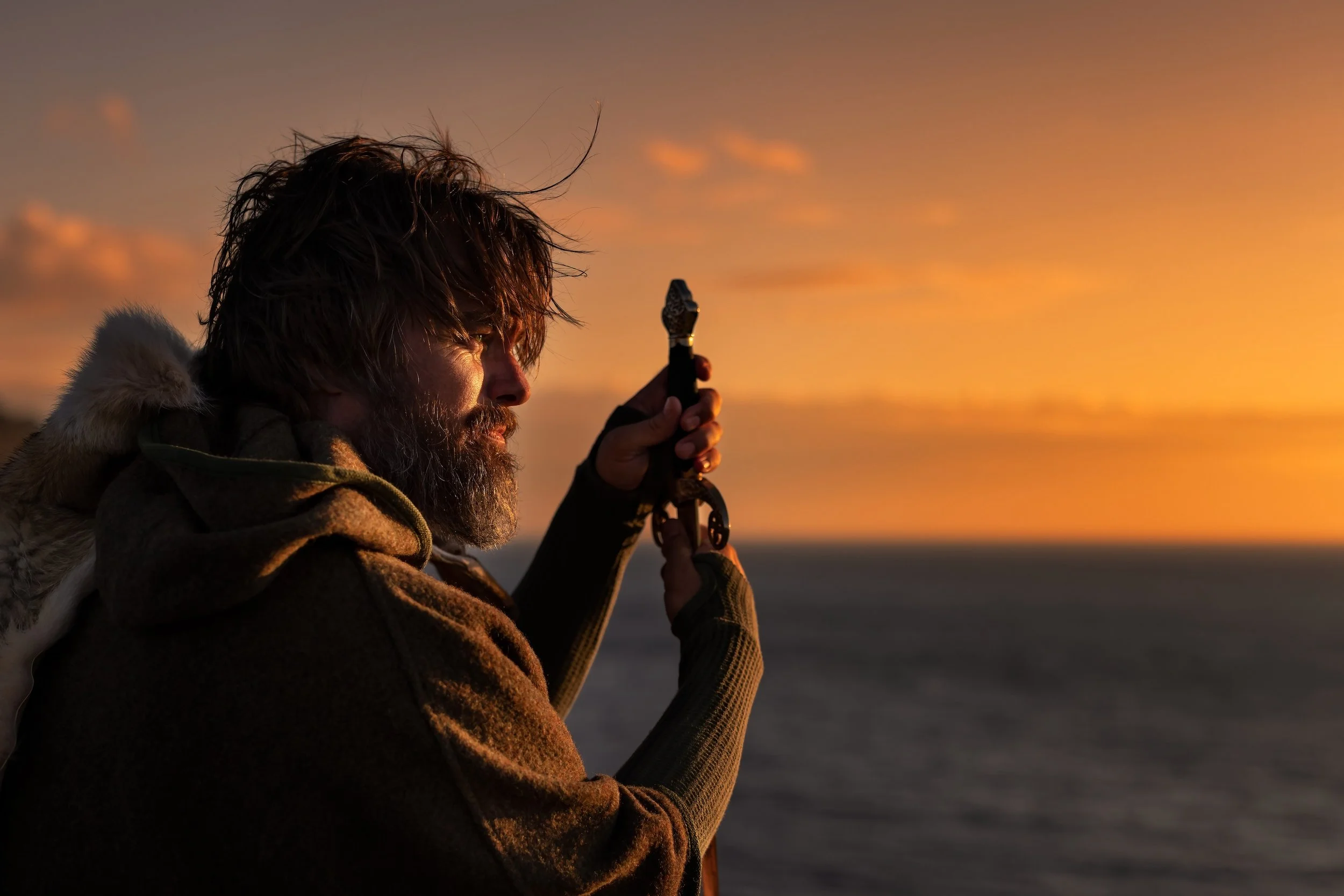 A man with long hair and a beard, wearing a brown jacket with a fur-lined hood, holding a fishing rod and looking at the sunset over a body of water.