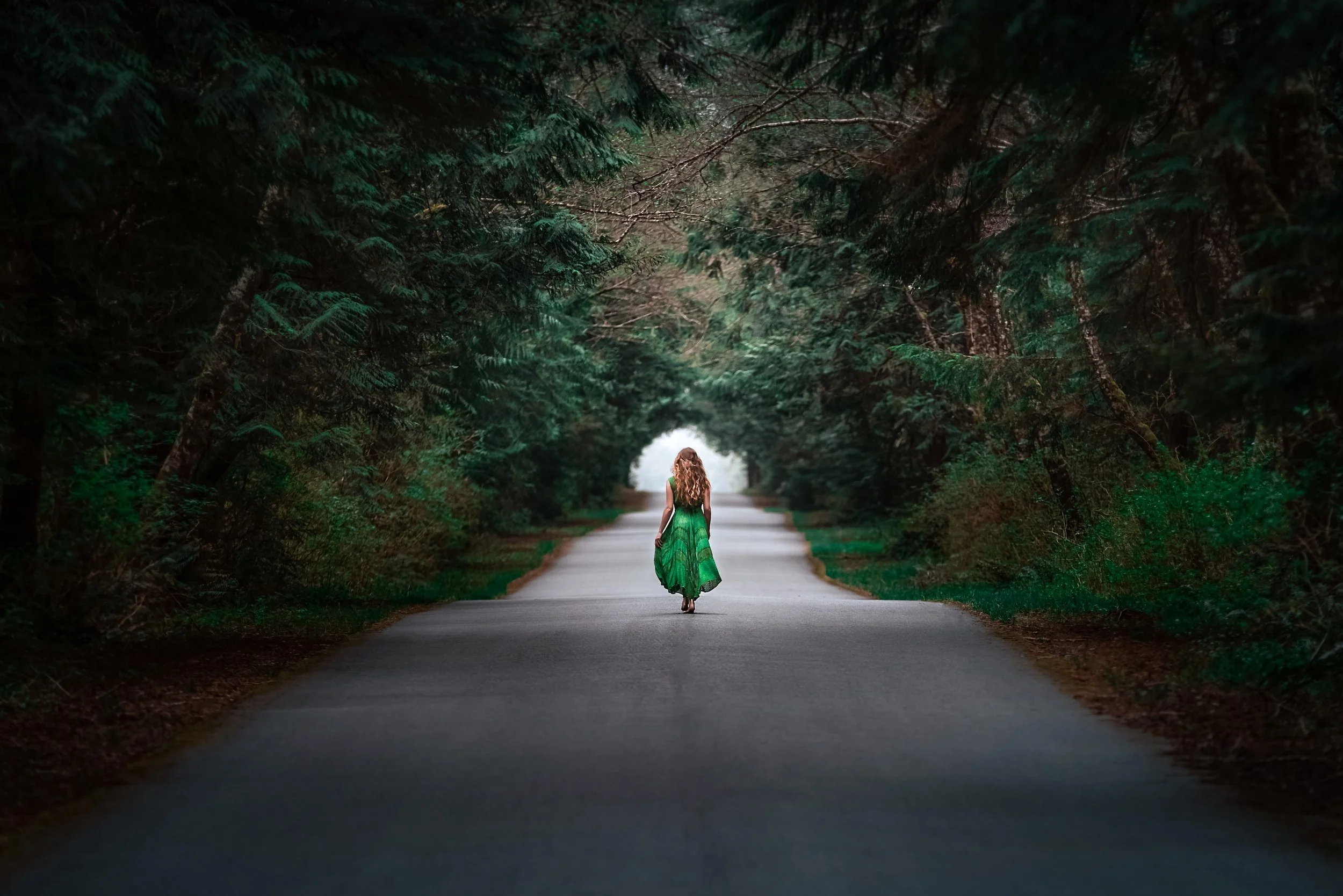 A woman with long, wavy hair wearing a green dress walking down a forested road surrounded by tall trees and greenery, with light at the end of the road.