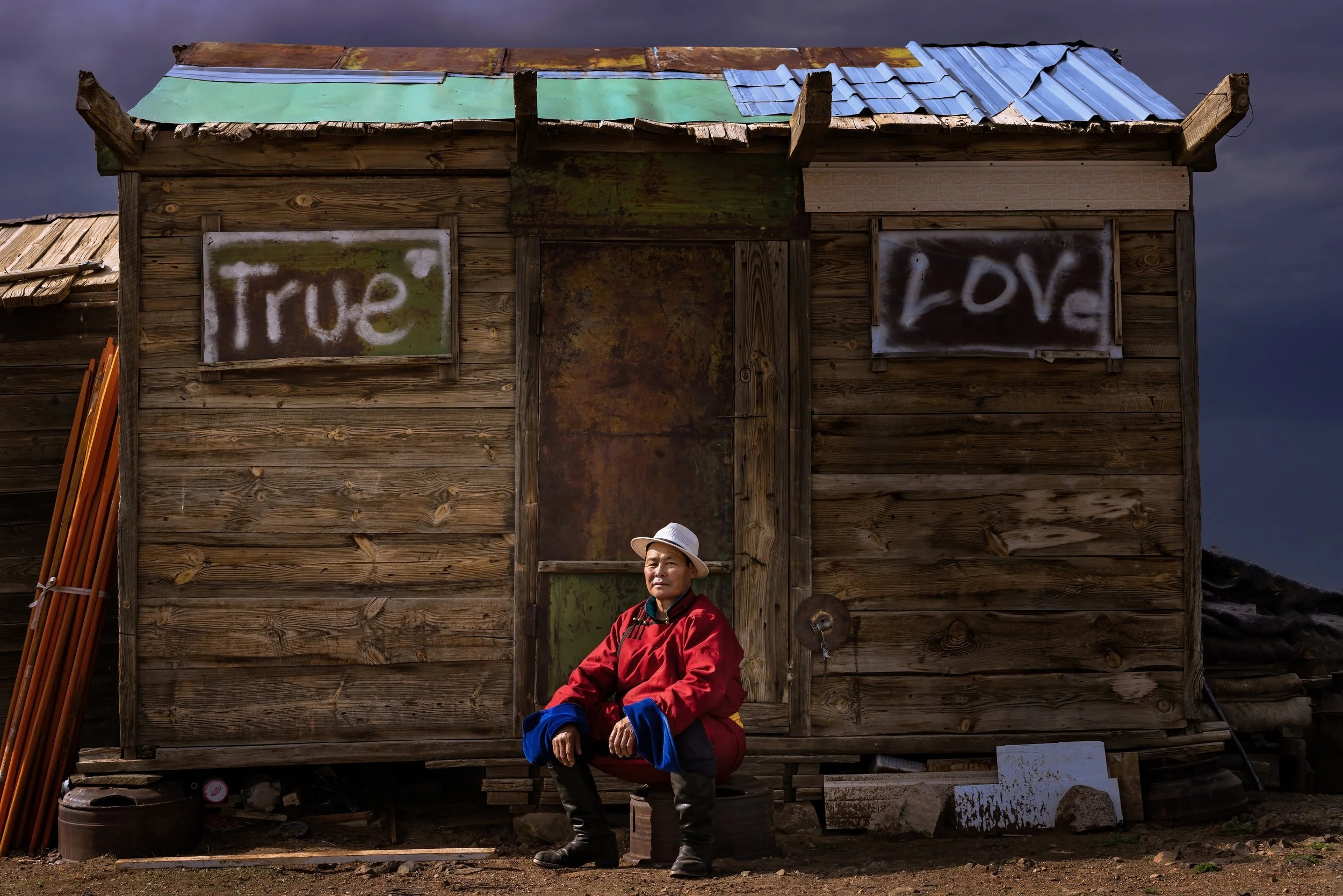 A person wearing a red jacket, black boots, and a white hat sitting in front of a weathered wooden house with spray-painted signs that read 'True' and 'Love'; dark clouds in the sky.