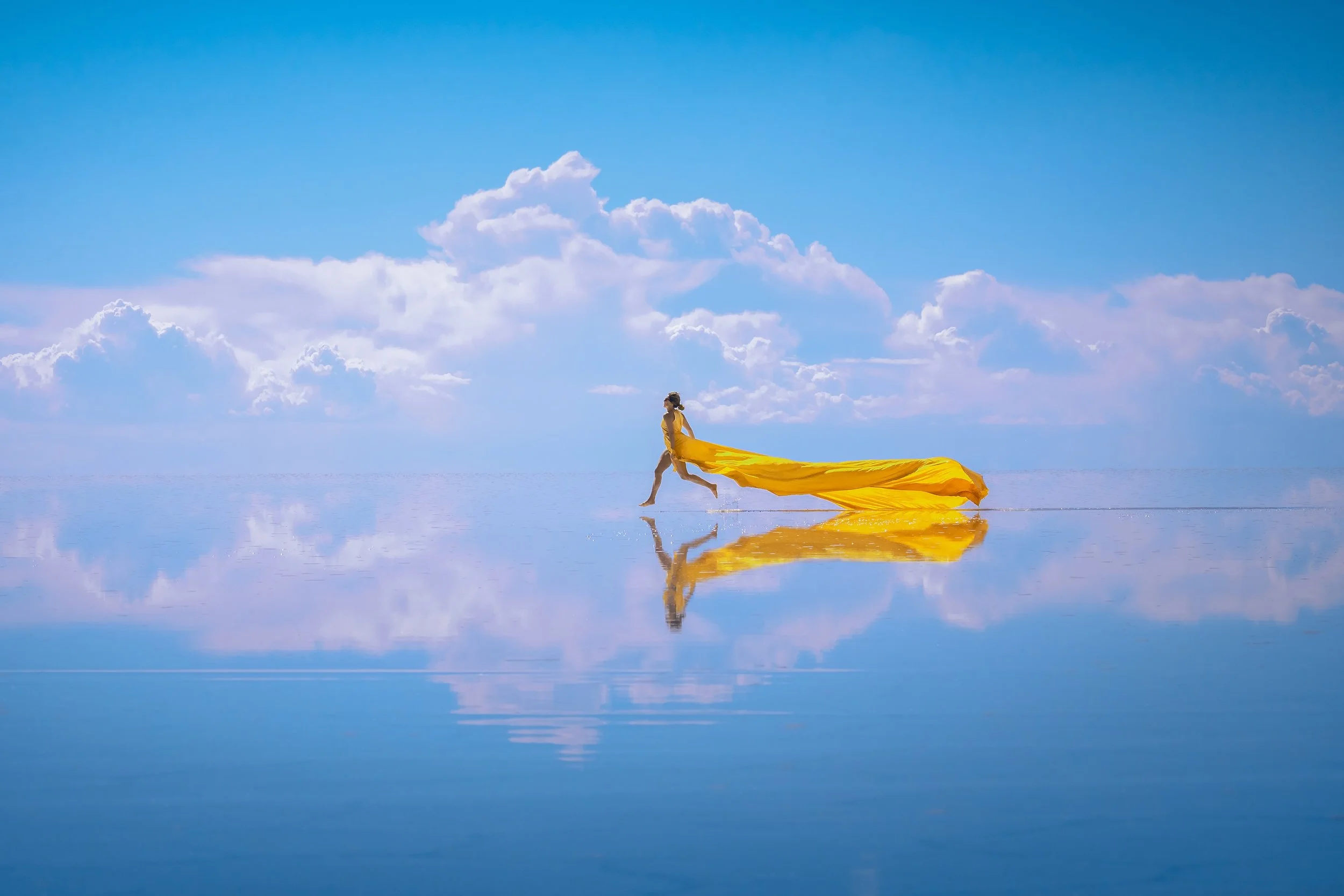 A person in a white swimsuit running on a reflective surface beneath a blue sky with clouds, holding a flowing yellow fabric, creating a mirror reflection in the water.
