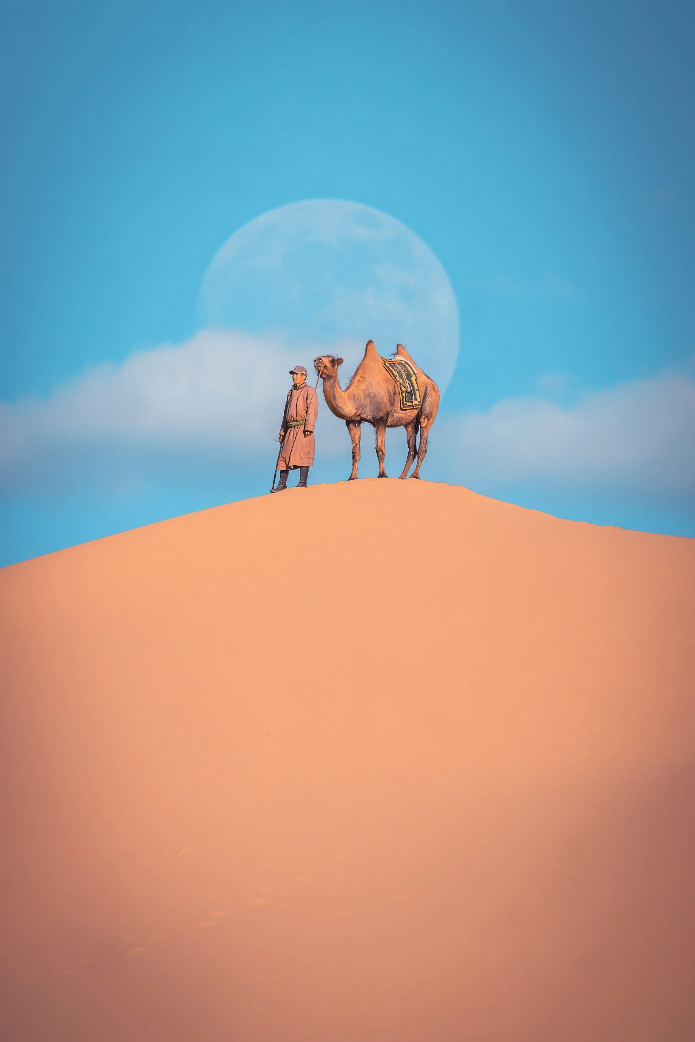 A person standing beside a camel on a sand dune with a large full moon in the sky in the background.