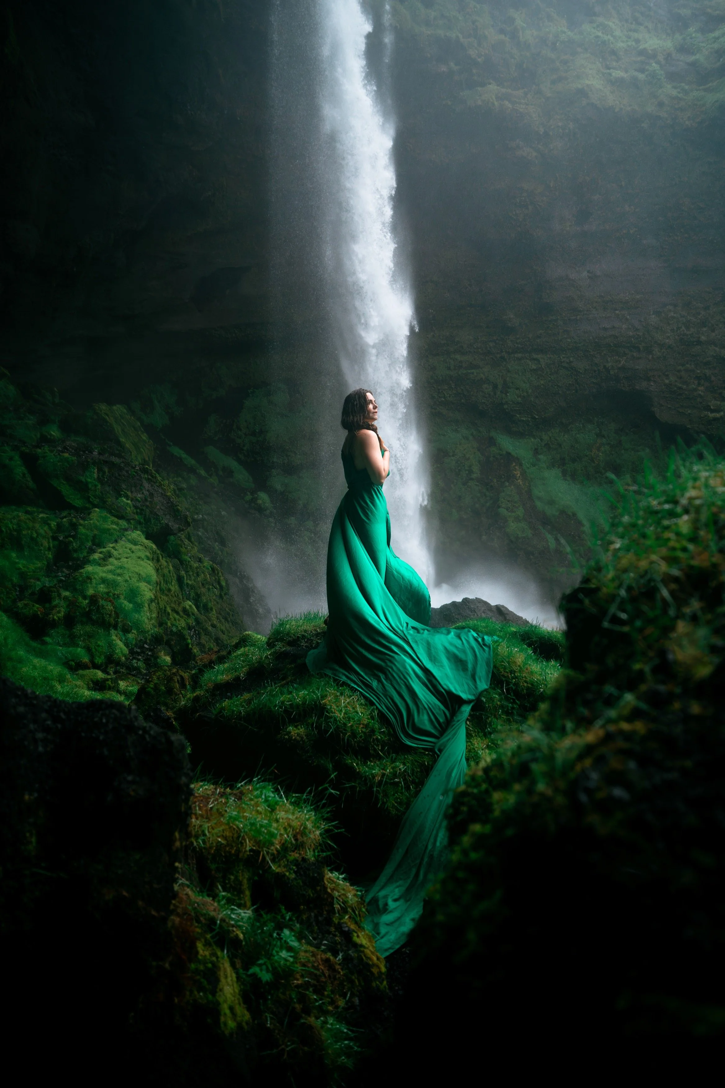 A woman in a green flowing dress standing on mossy rocks near a waterfall in a lush, green forest