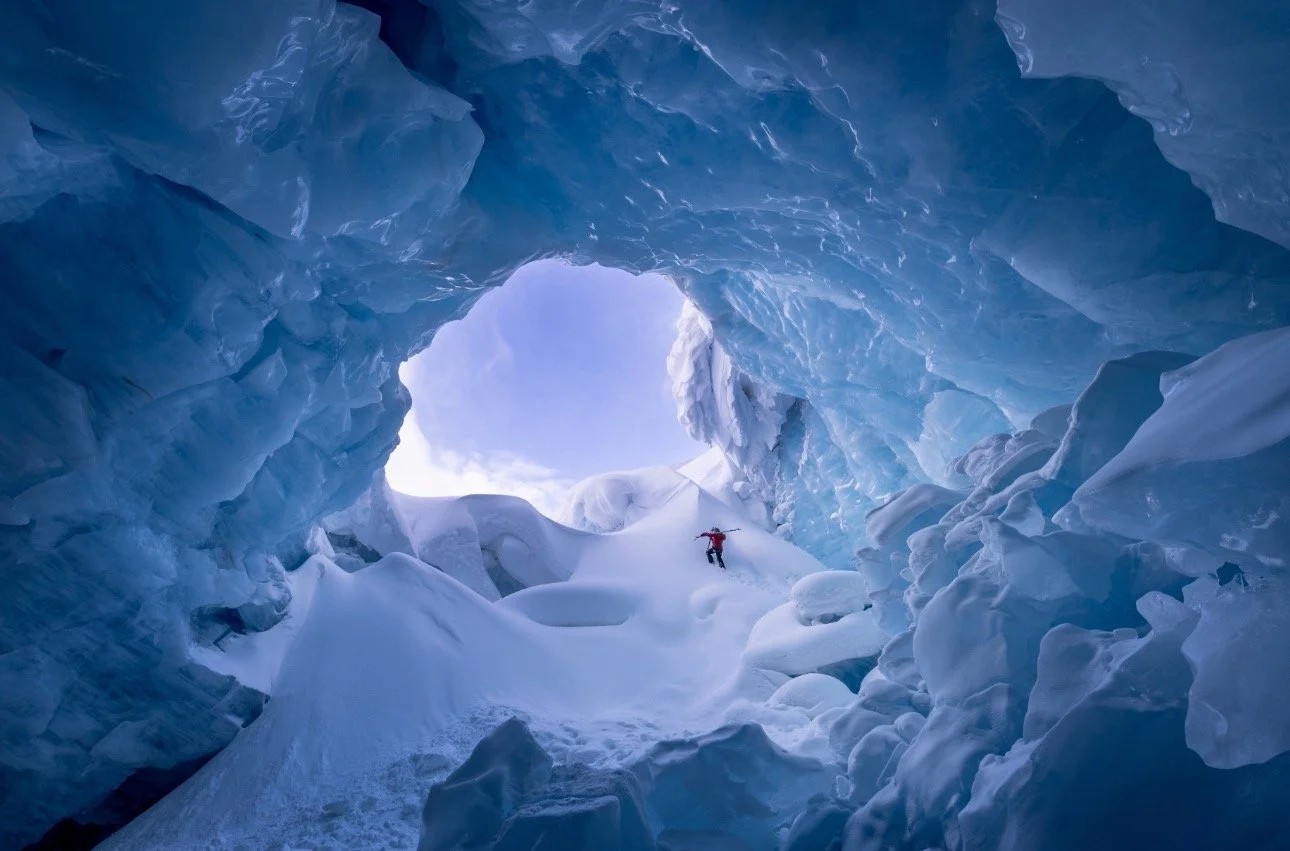 A person in red gear climbing inside an icy blue glacier cave.