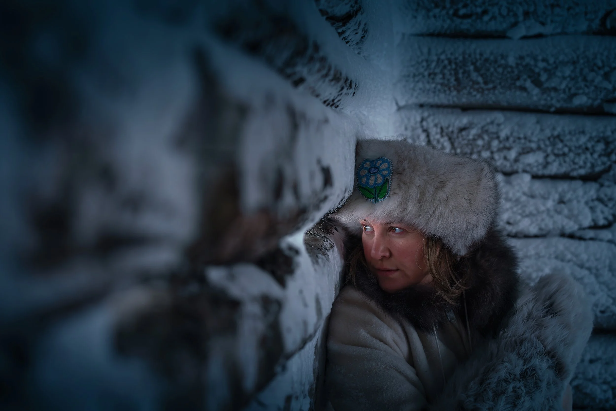 Person wearing fur hat with embroidered blue flower, leaning against a snowy stone wall in a cold environment.