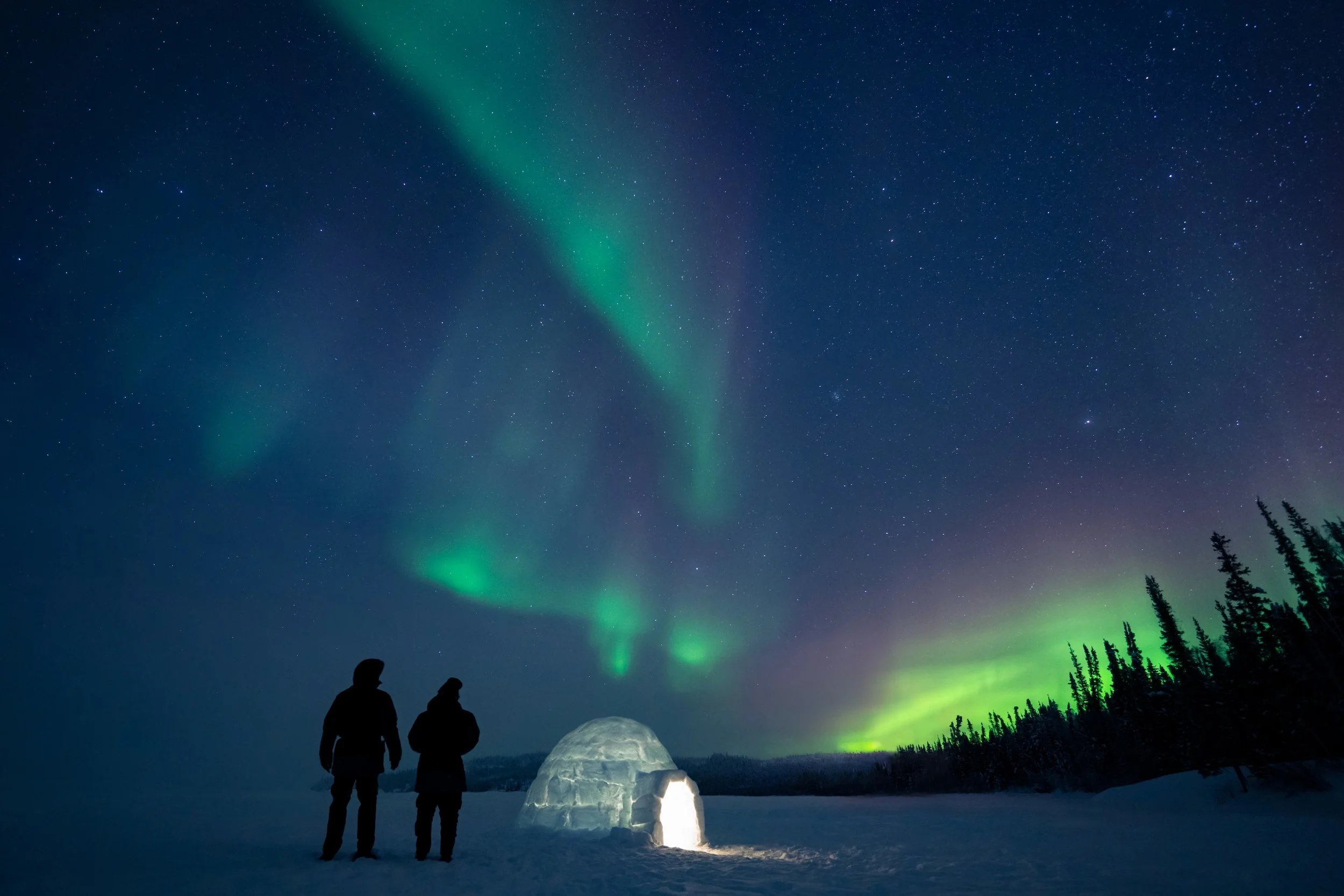 Two people standing by an igloo in a snowy landscape at night, watching the Northern Lights in the starry sky.