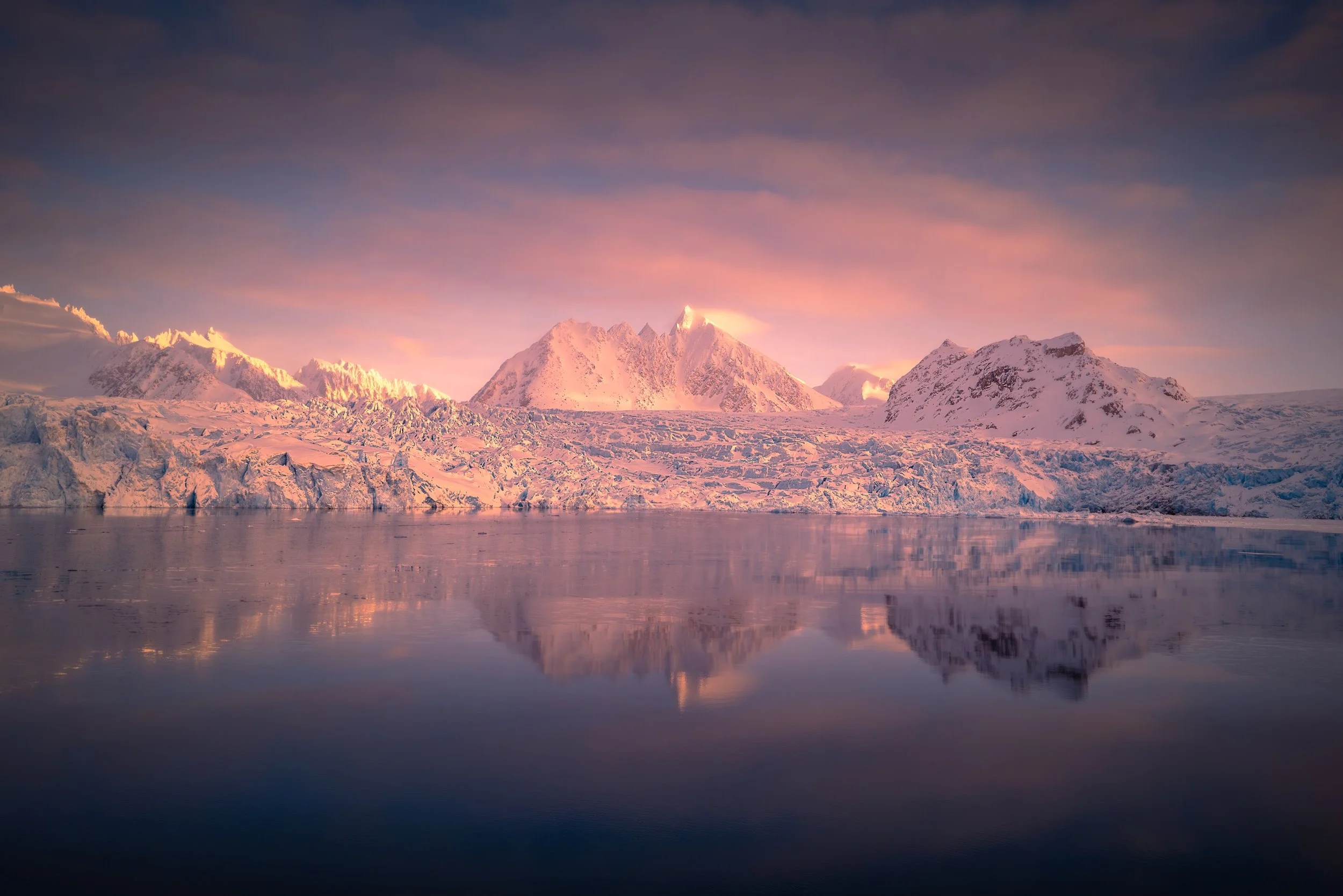 Snow-capped mountains reflected in calm water during sunset with pink and purple hues in the sky.